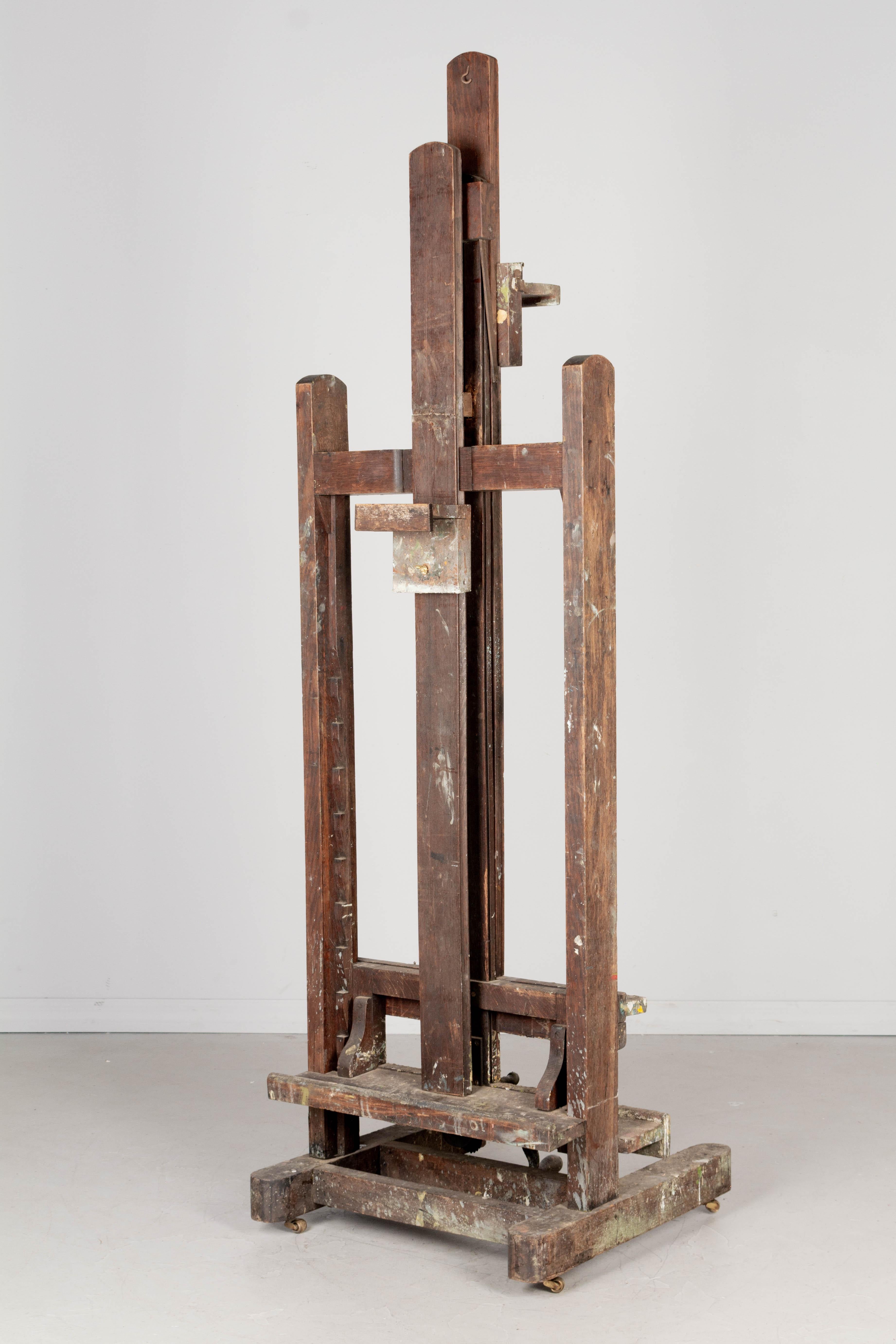 An early 20th century French artist painter's easel made of solid oak with thick layers of old paint, iron threaded vise and wood handled crank. Double sided with large deep ledges and adjustable clamps to securely hold the painting in place with