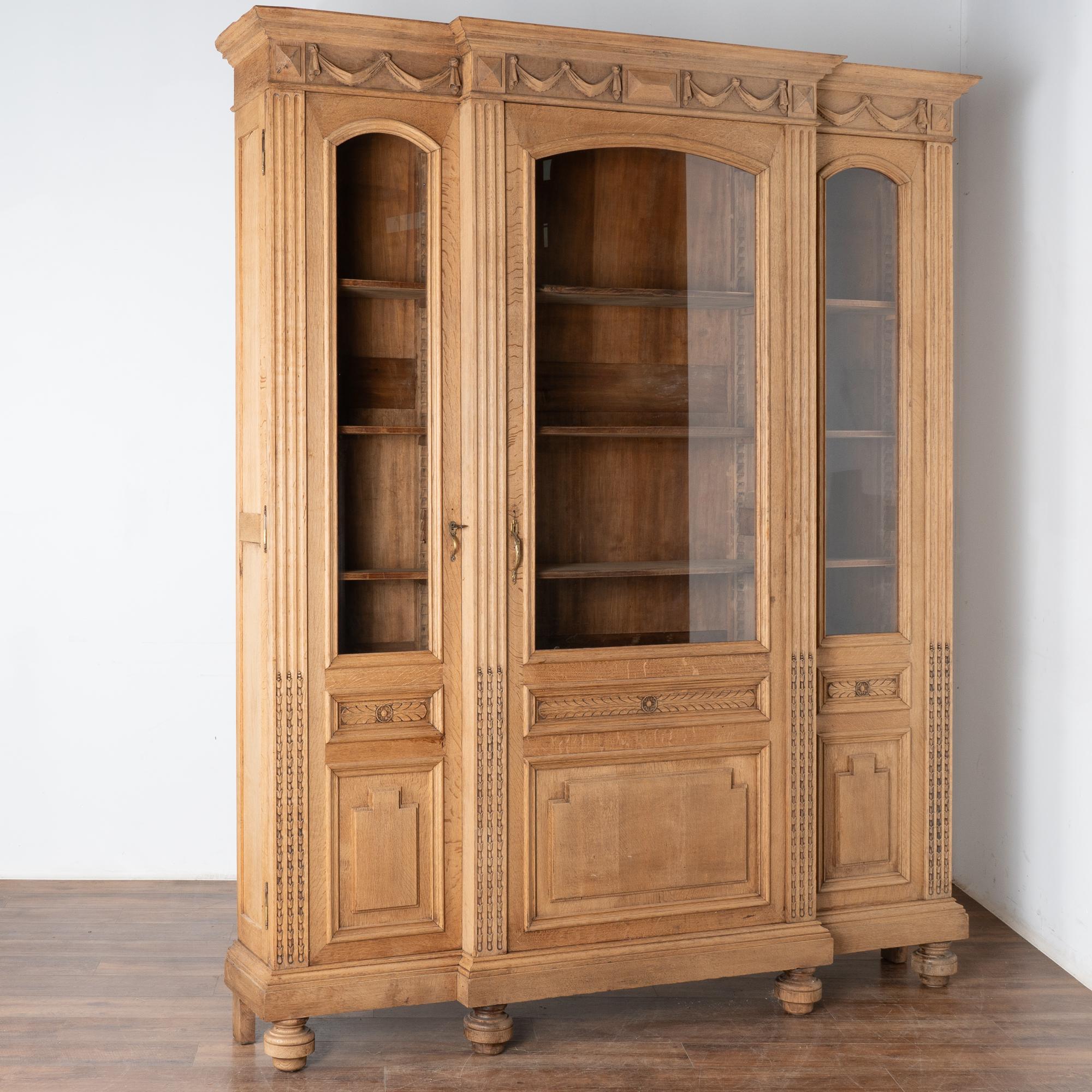 This large bleached oak bookcase or display cabinet has three long glass panels with heavy carved panels on bottom and stands over 8' tall.
Ideal for display of books or any collection, the interior shelves are adjustable (pegs included).
Notice the