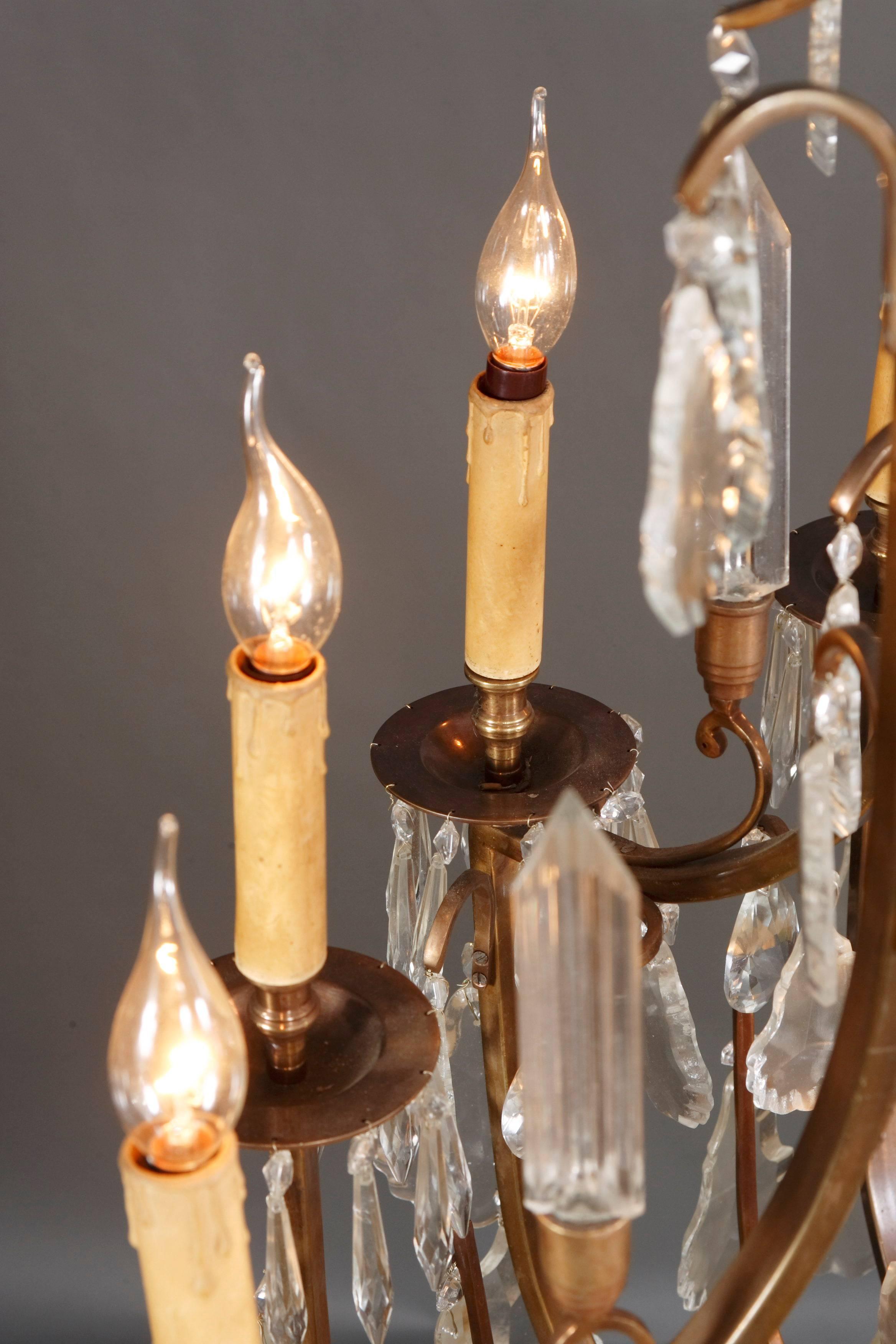 High pear-shaped body. Multi-curved Korbgestänge. In the center ascending, multiple articulated baluster-shaped glass shaft with multiple crowning. Brass, chased. Twelve outboard flames and six polished glass obelisks. Candle-shaped pods made of