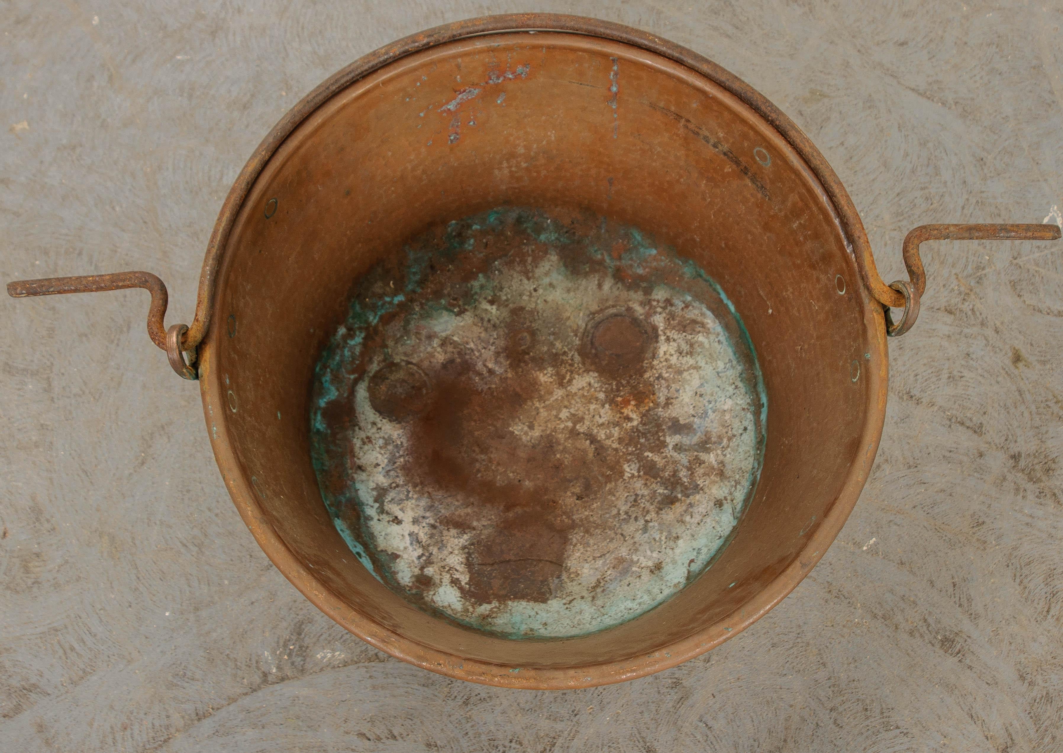 A sizeable antique copper pot, equipped with a forged iron handle that can be used to suspend the vessel over a fire. The pot has a hammered exterior that gives the antique a styled finish. The copper has patinated wonderfully, with some verdigris