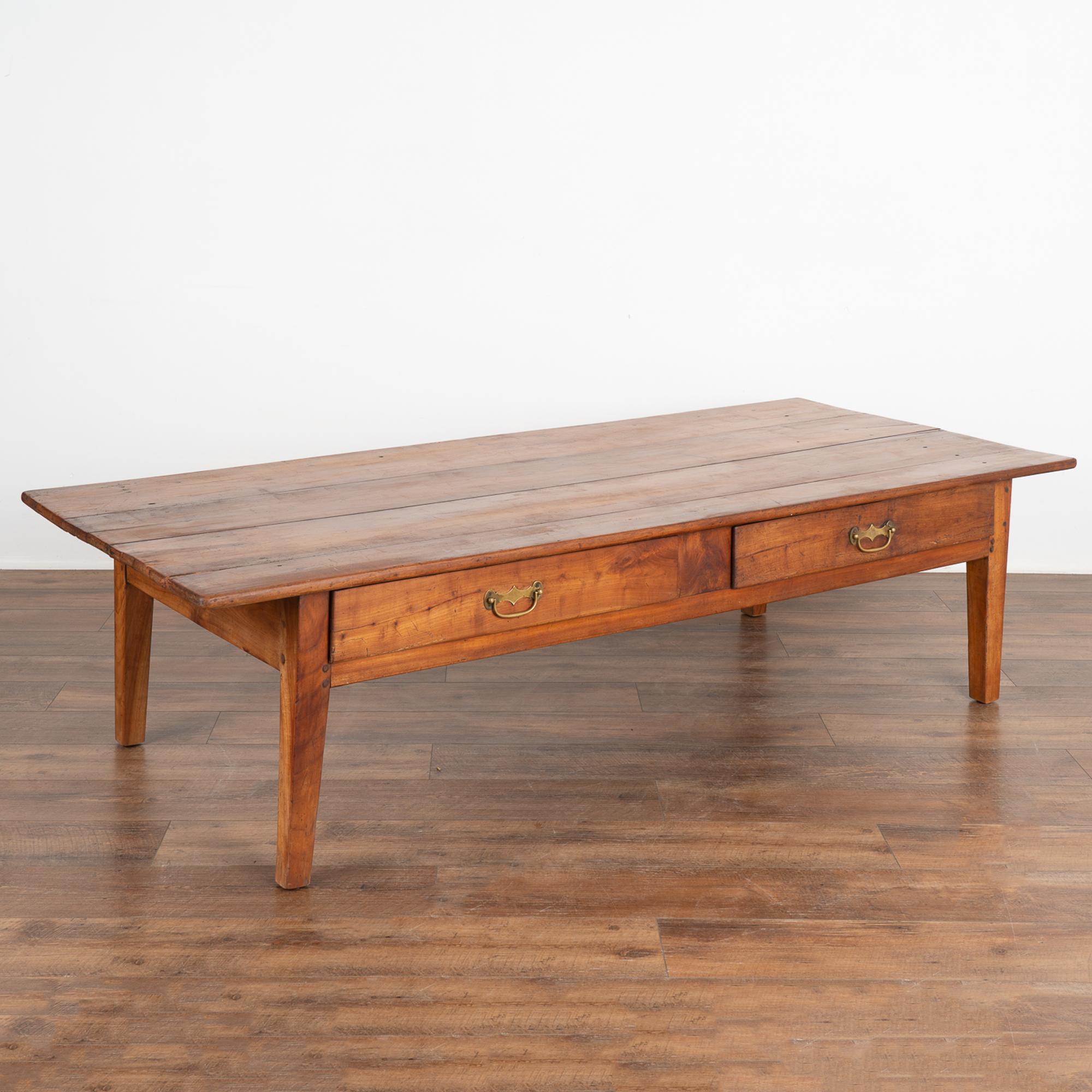 The beauty of this large French country coffee table comes from the warm aged patina of the cherry wood. The top is made from four planks, and skirt holds two functional drawers with brass pulls. 
Restored, strong and stable. Any scratches, cracks,