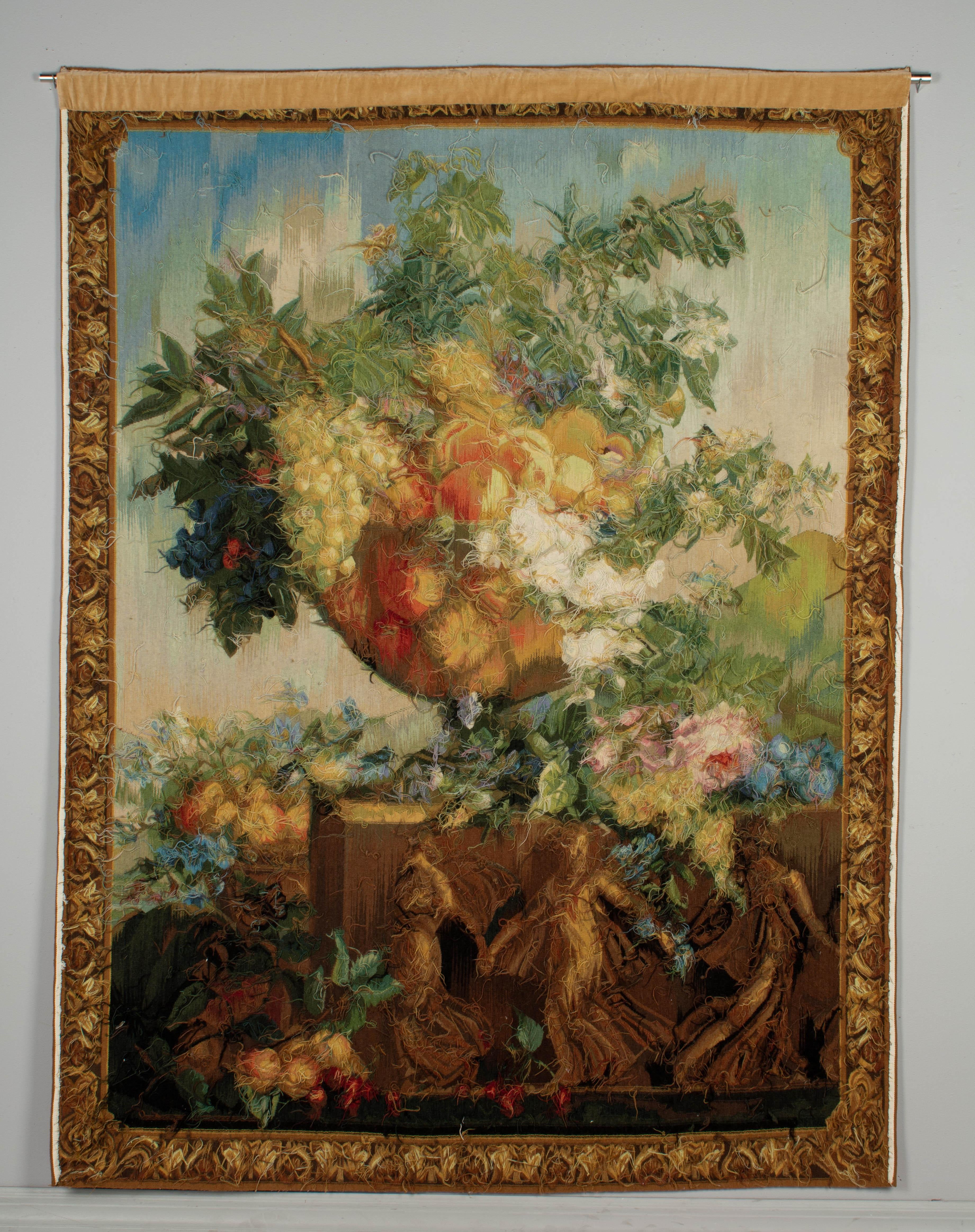 A large French wool tapestry, or wall hanging, depicting a Nature Morte, or floral still life. A large garden urn overflowing with flowers and fruits, including grapes, peaches, plums cherries and large pink roses. The urn rests on a large plinth
