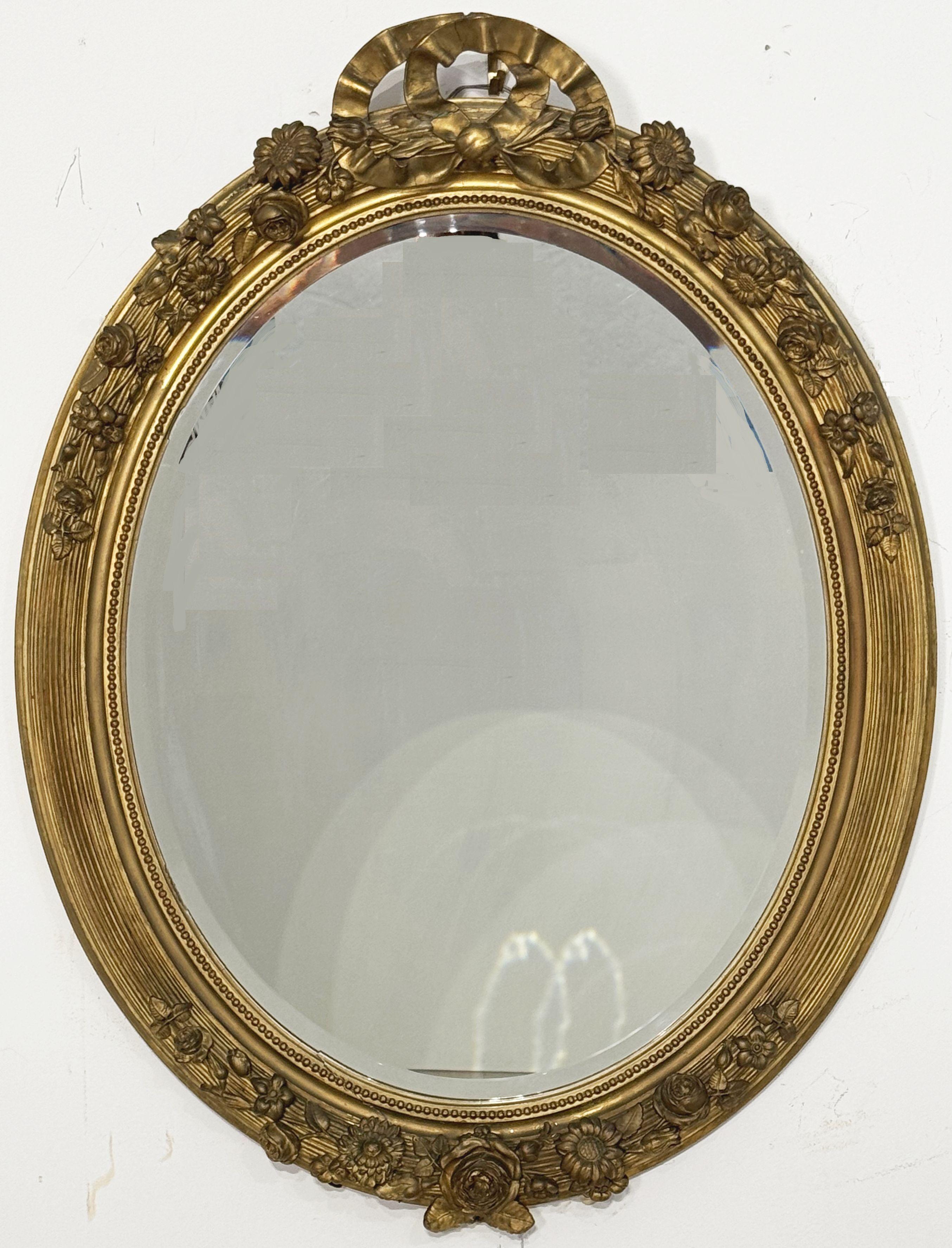 Gran espejo francés dorado de pared o de recibidor, con marco oval en relieve, cresta floral con lazo y friso inferior -a la manera de Luis XVI, con estilo rococó o barroco- que rodea una placa de espejo biselada en el centro.

Dimensiones totales: