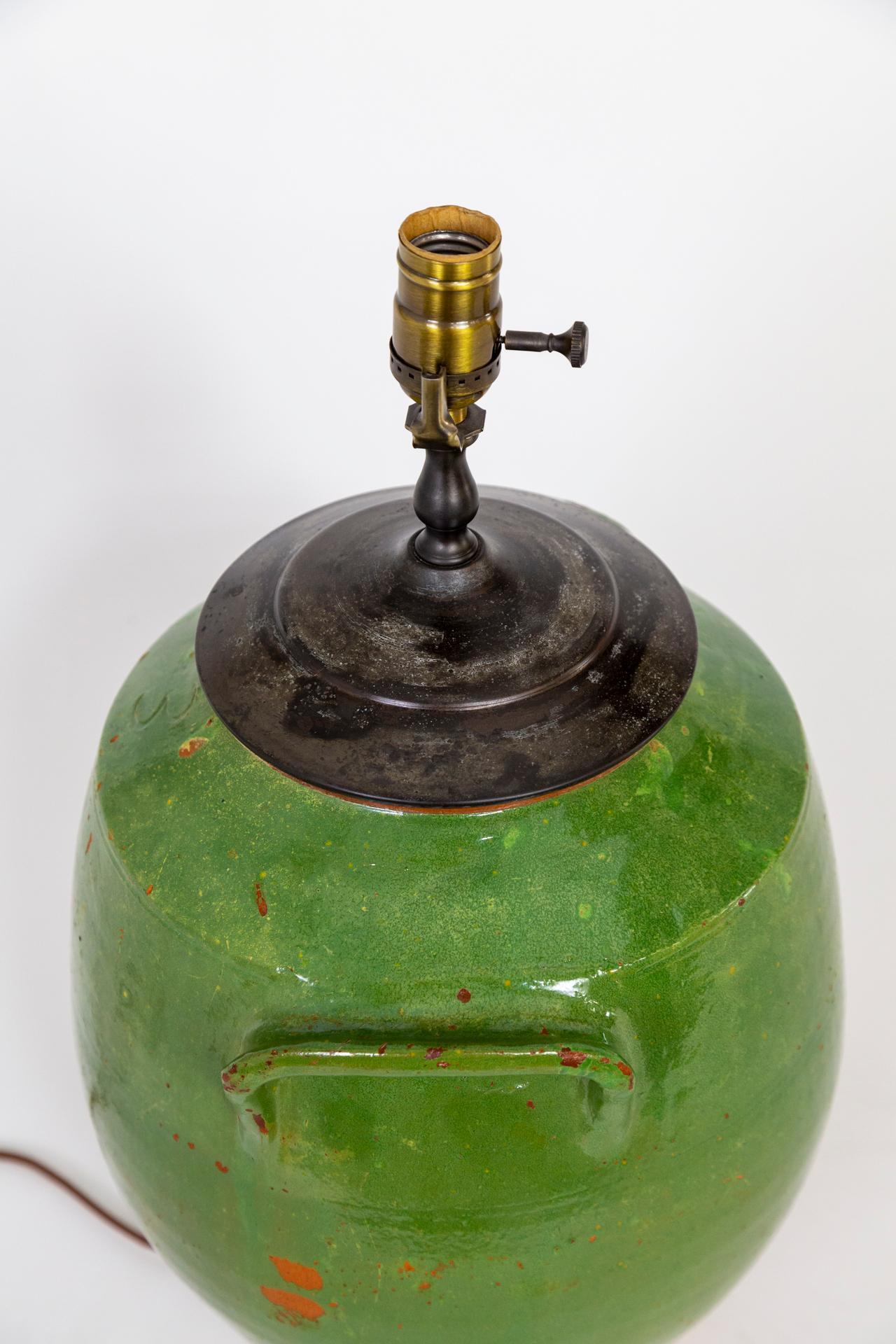 A French, green glazed, earthenware jar mounted as a table lamp. Beautiful, deep gray, speckled patina on bronze top cap, with a wood base matching the terra cotta at the bottom of the form. 3-way socket. 21” to top of socket x 12” width x 10” depth.