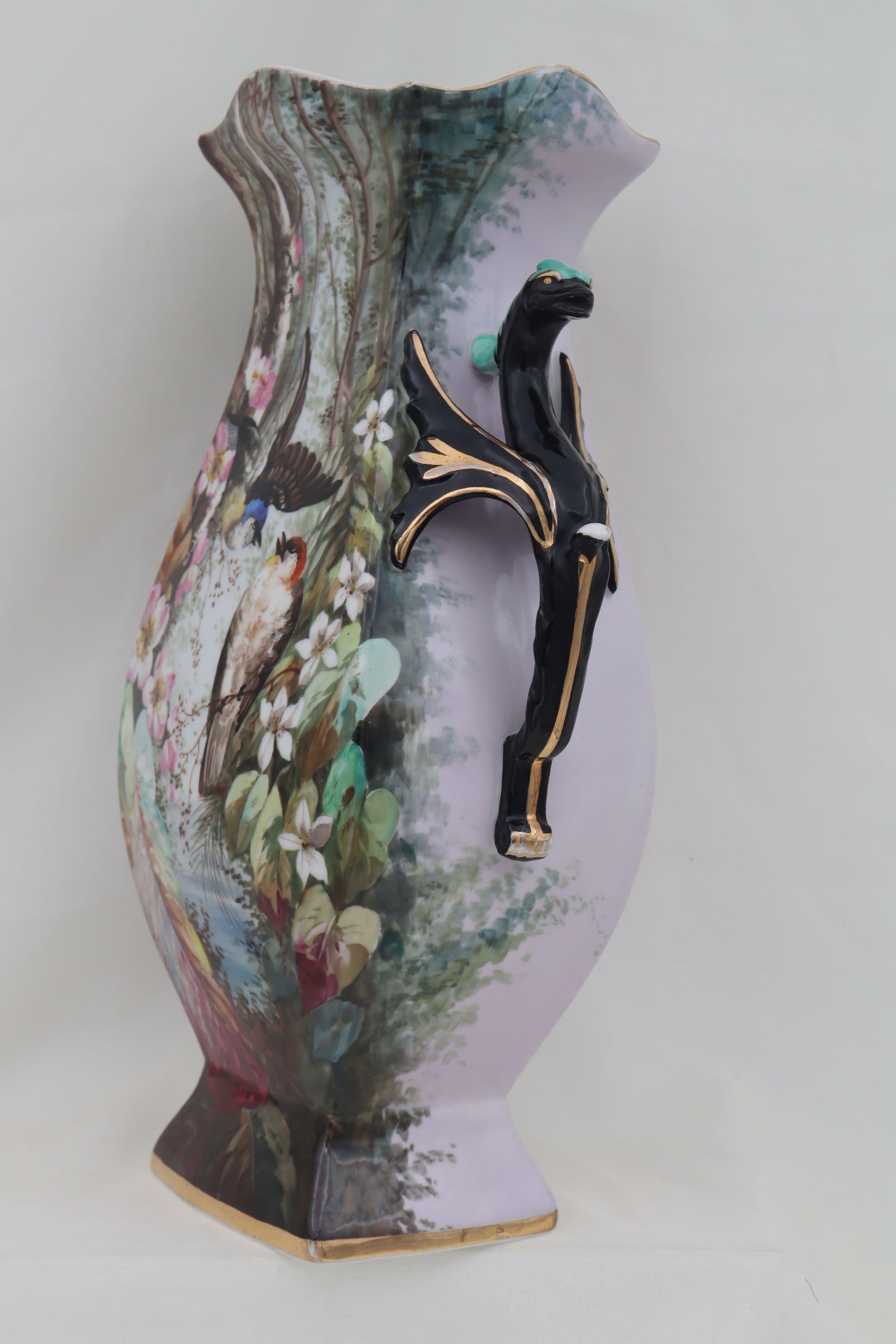 This large French porcelain hand painted and gilded vase features two colourful birds perched on branches in a forest setting surrounded by blossoms and coloured leaves, on a pale lilac ground. The handles are mythical winged serpents and their