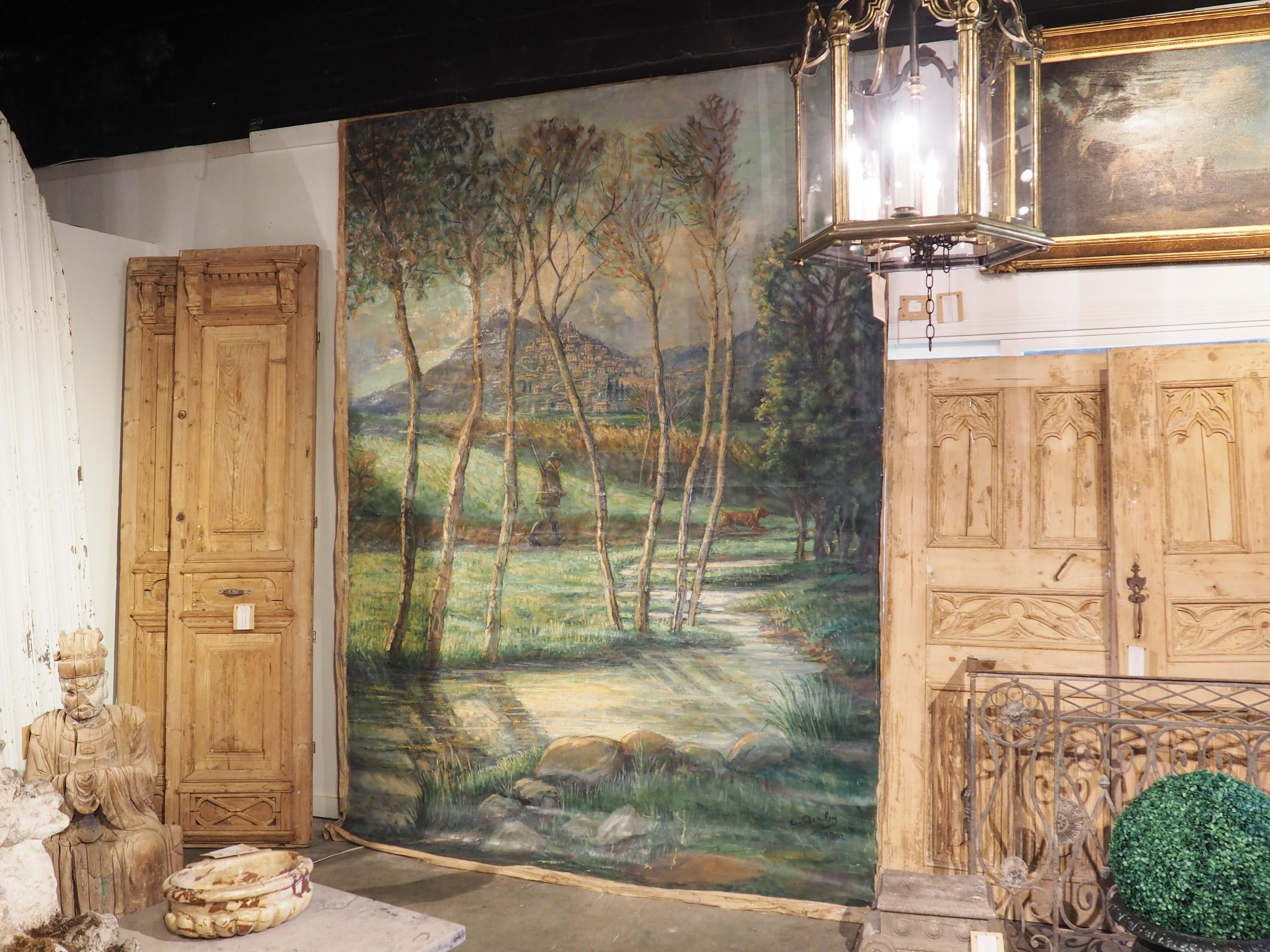 Purchased from a chateau in the South of France, this large landscape painting of a hunter walking outside of a hillside village is over 10 feet tall and seven feet wide. The canvas is signed and dated in the lower right-hand corner, indicating that