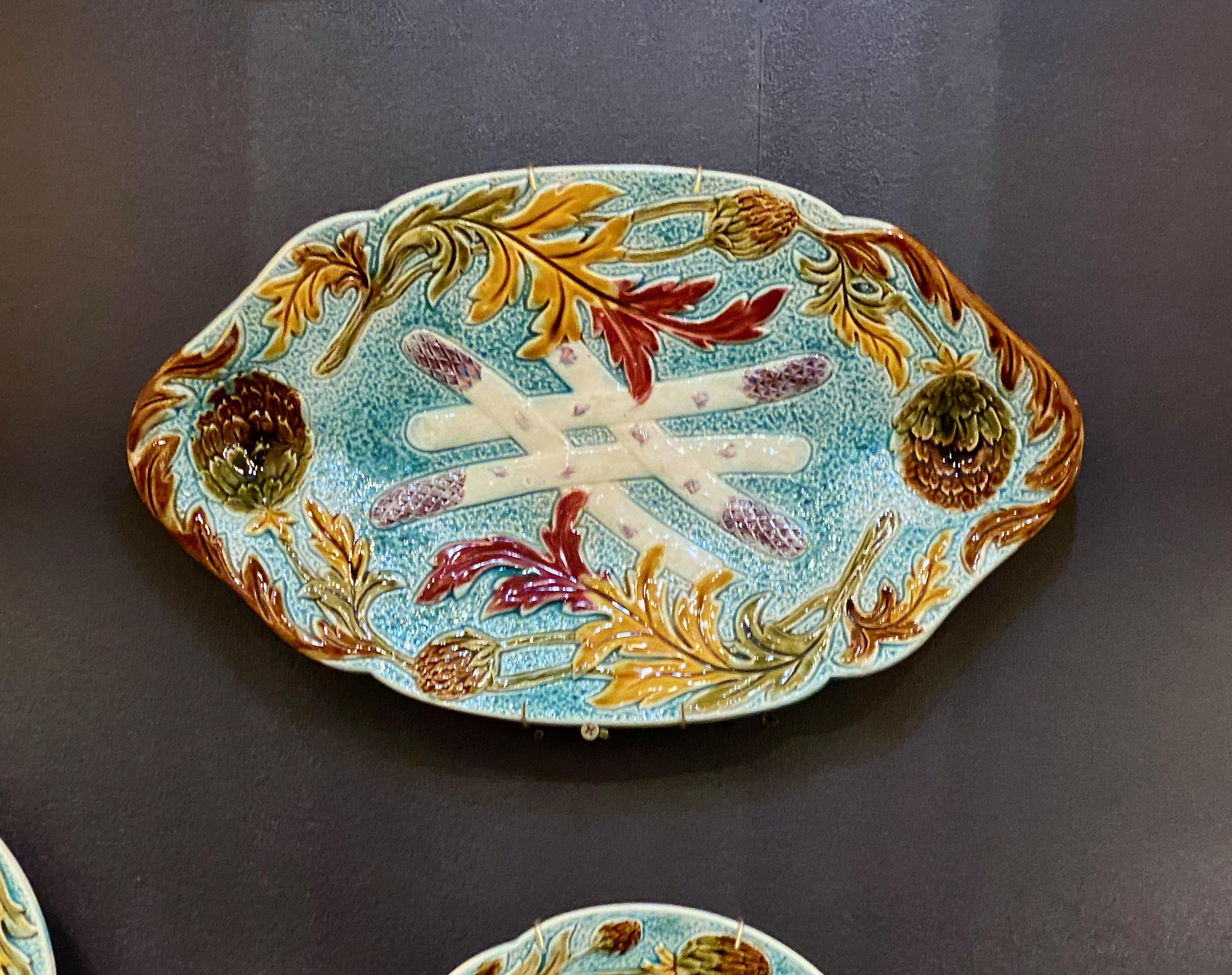 This is a large vibrantly toned asparagus platter in French majolica or faience that dates to c. 1880. The platter features asparagus spears in relief together with artichokes and scrolling leaves. There is a sauce well in the form of an