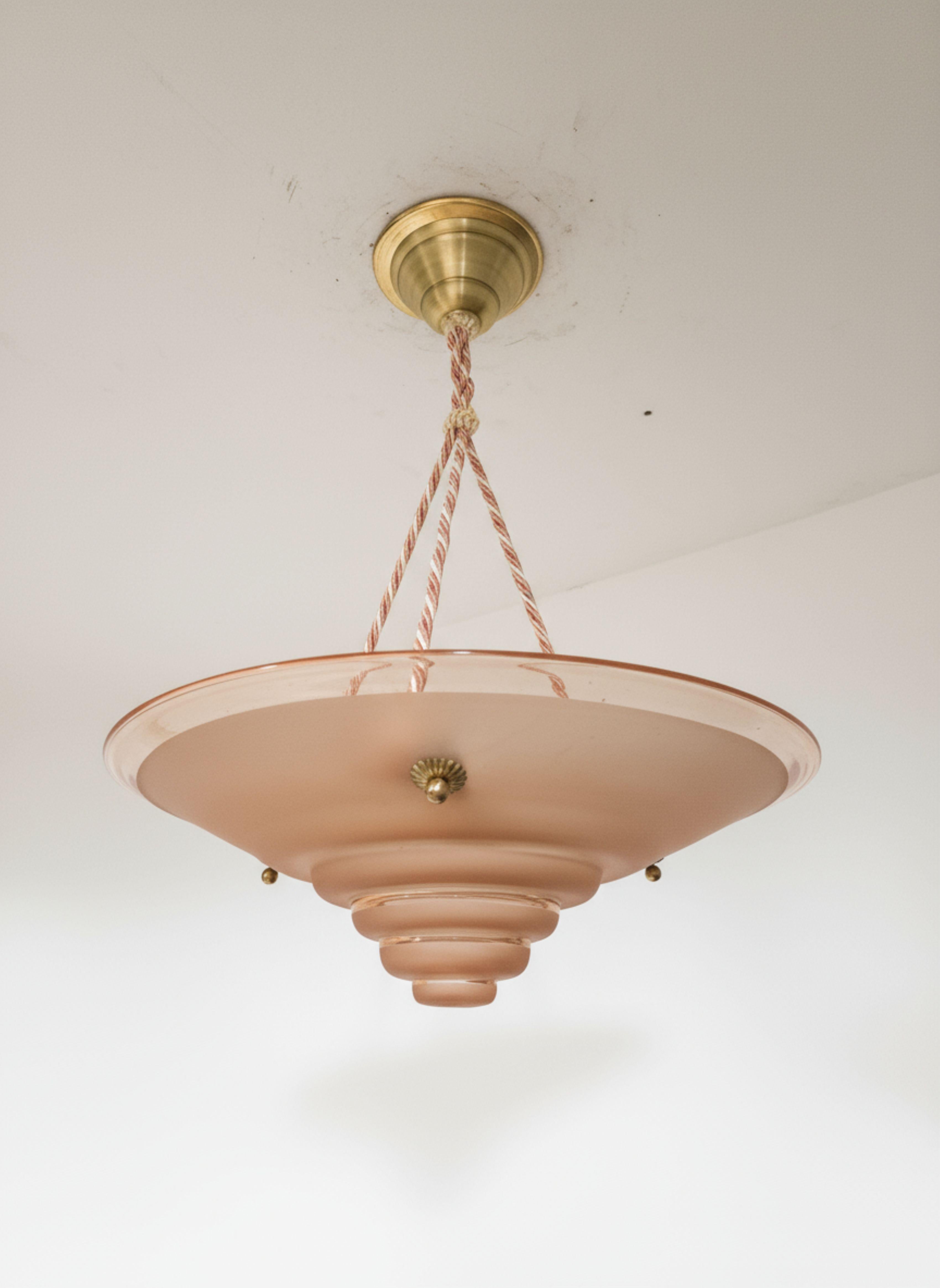 An elegant Art Deco pendant ceiling lamp featuring a distinctive stepped art glass glass shade in a soft pink hue.   The fixture is hung by a pink and beige rope twist cord, which complements the period design. Finished with a polished brass canopy
