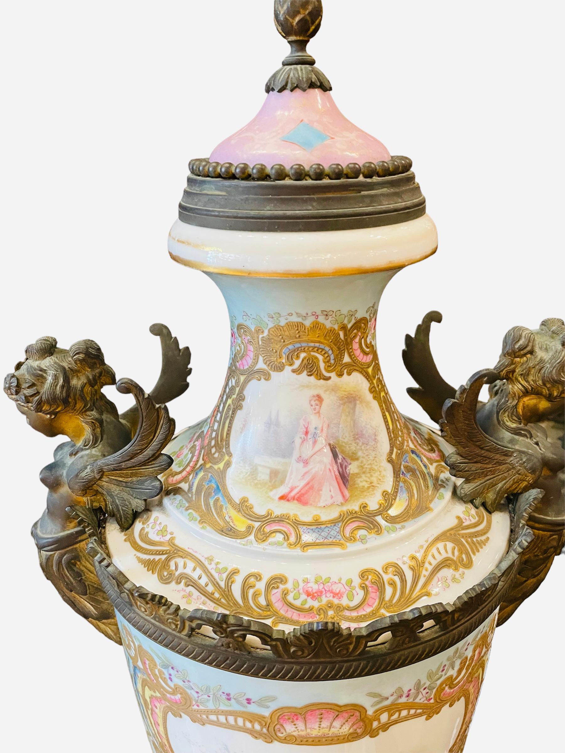 This is a large and heavy French Sevres style patinated bronze mounted porcelain lidded urn. It is hand painted in a white and sky blue background. It depicts a scene of an 18th century gentleman greeting a lady in the front center body of the urn.