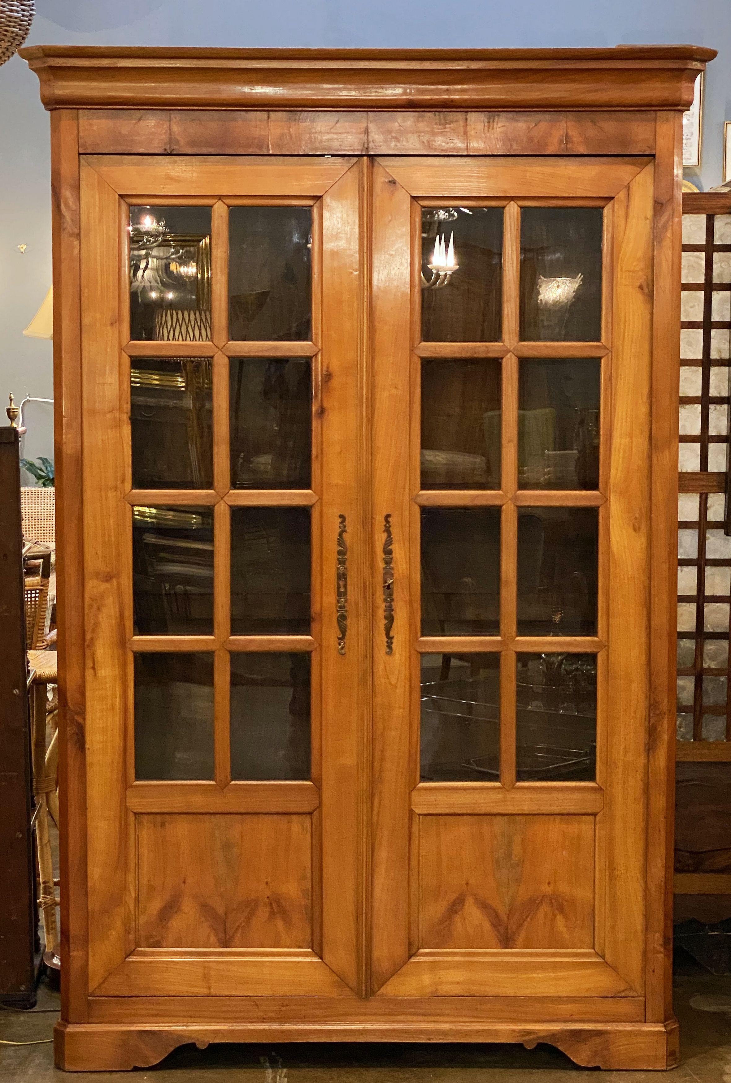A handsome large French bookcase cabinet of cherry from the 19th c., featuring a moulded crown top over a frieze of two glazed paneled doors, the doors with decorative hardware and escutcheon, opening to an interior with four shelves, and standing