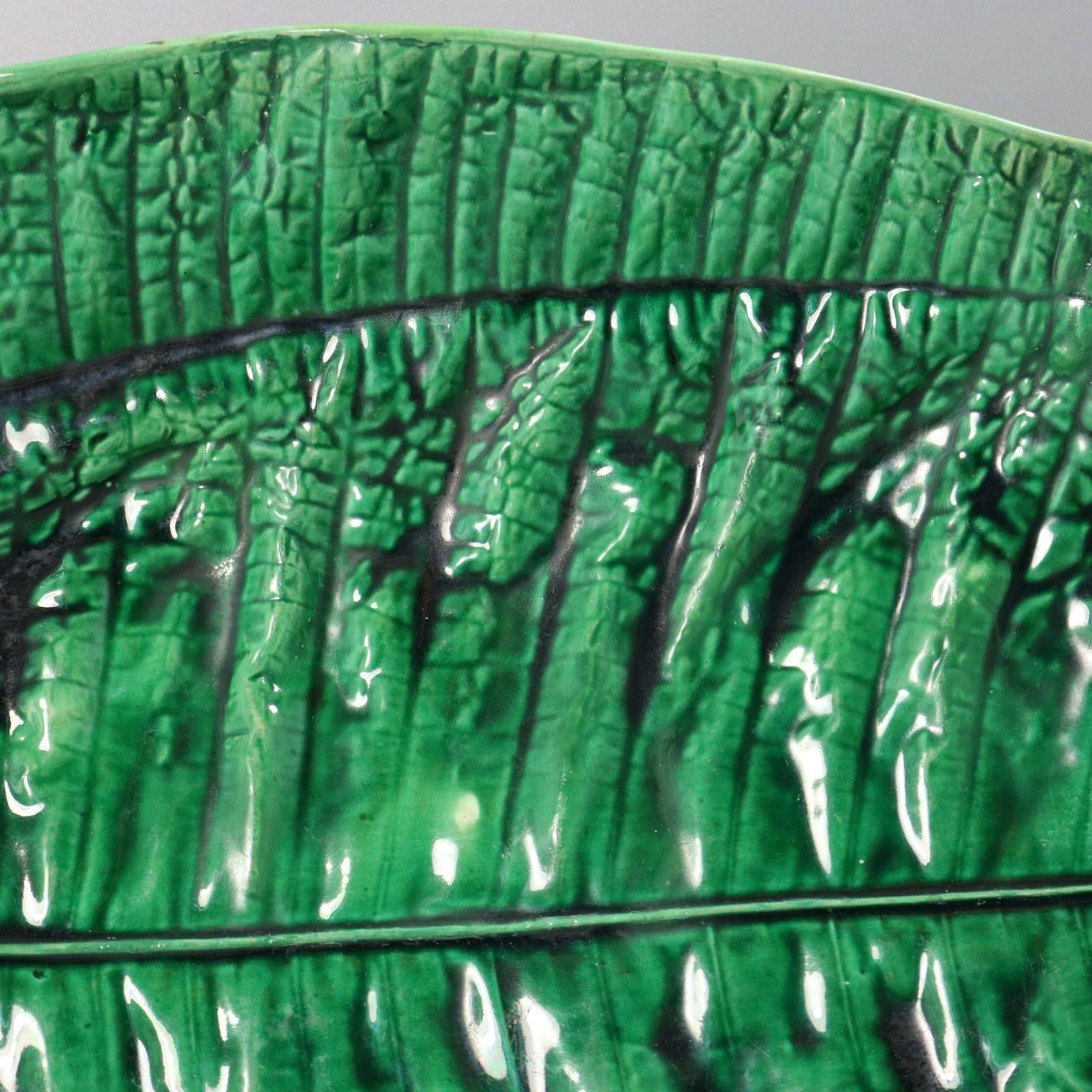 Plat en majolique de George Jones représentant une feuille de bananier sur une base en forme de branche. Coloration : le vert et le brun sont prédominants. La pièce porte les marques du fabricant de la poterie George Jones. Porte un numéro de