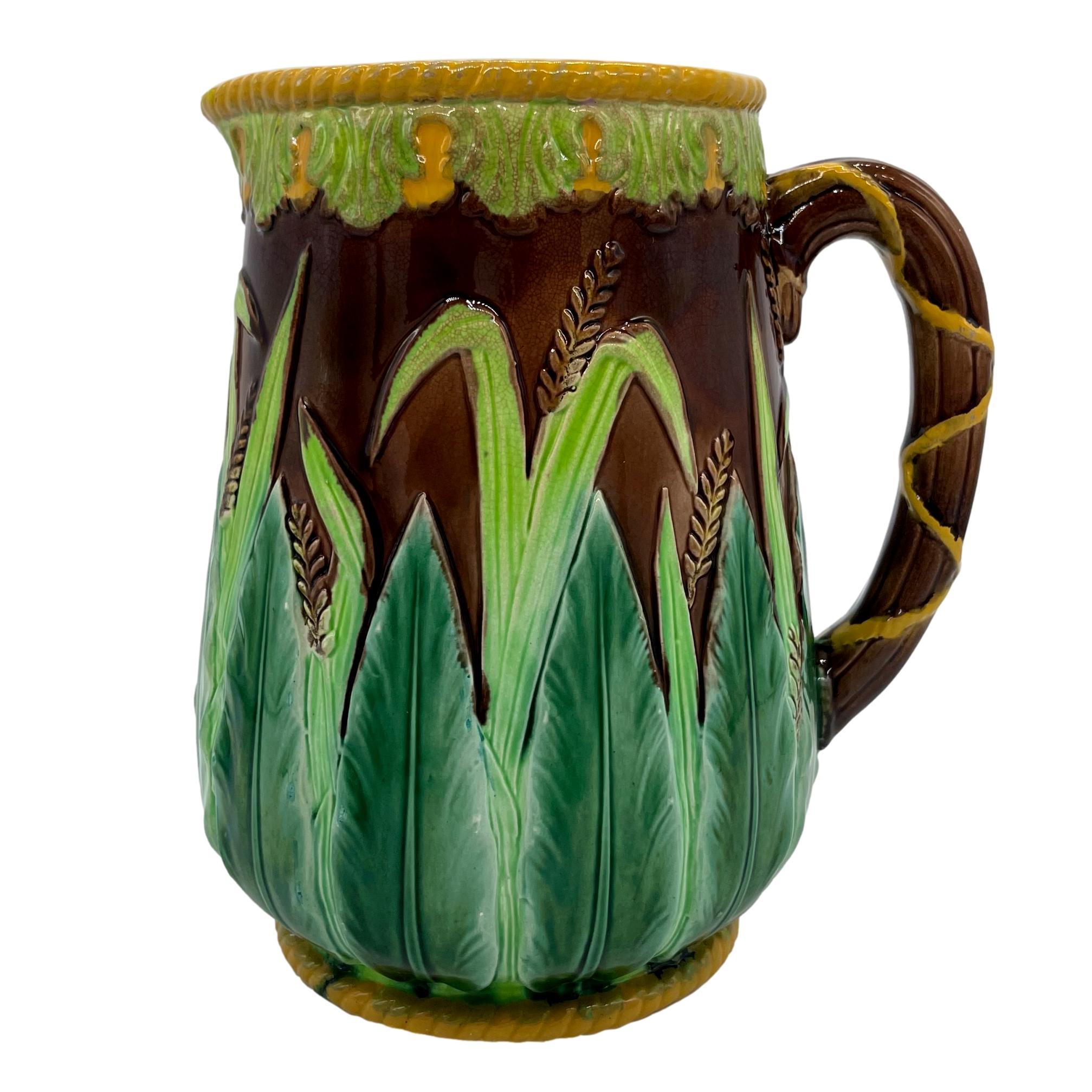 George Jones Majolica Table Pitcher, of baluster form with relief molded stiff acanthus leaves, grasses, and wheat on a brown ground, the top banded with stylized leaves glazed in chartreuse, with yellow glazed roping to the top rim and foot rim,