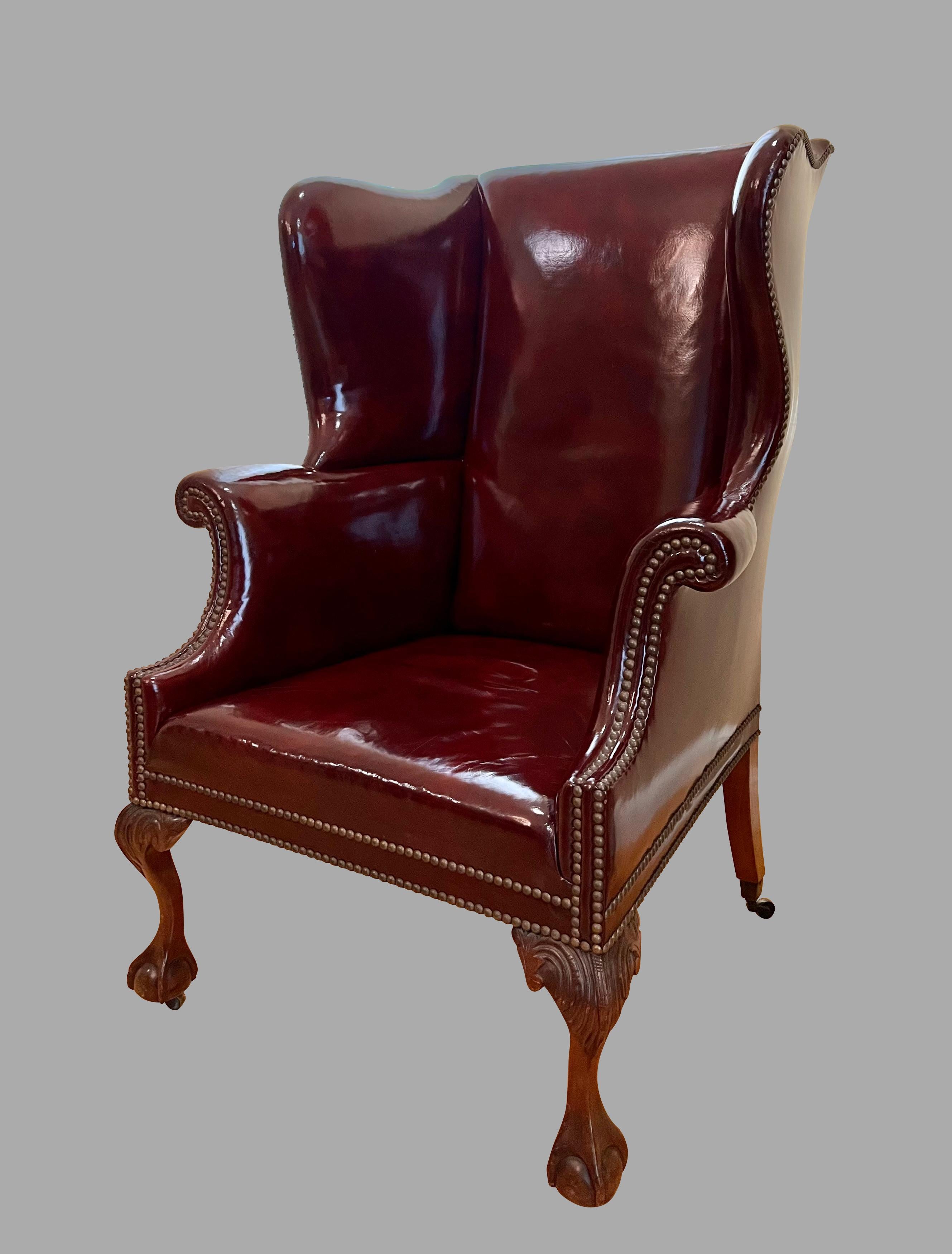 A good quality amply proportioned English Georgian style wing armchair of typical form, well-upholstered in polished claret colored hide finished with nailheads, supported on carved mahogany ball and claw feet ending in casters. Circa 1920.