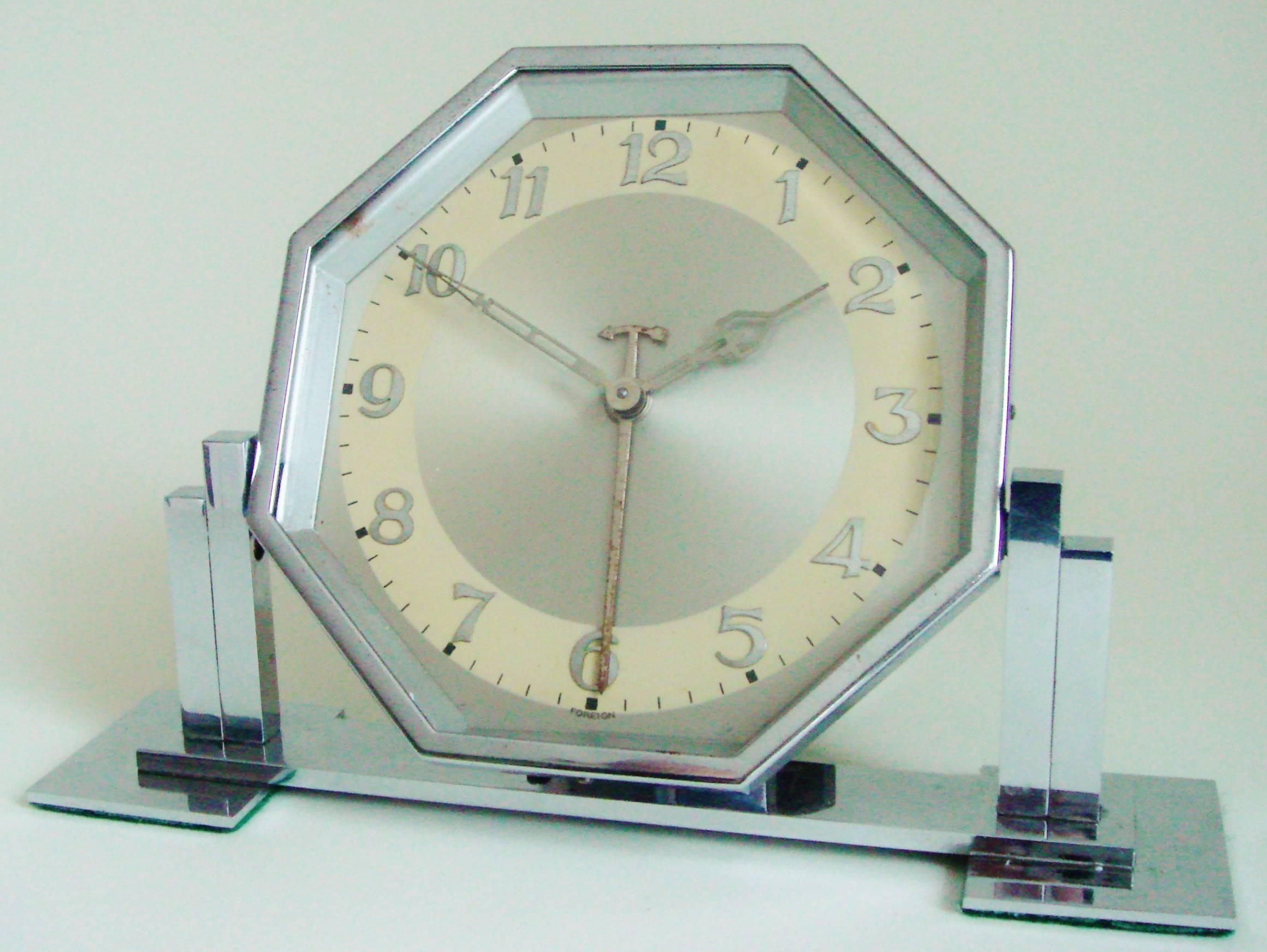This larger than usual German Art Deco chrome tilting alarm clock features a hexagonal body positioned between square twin stepped pillars of uneven height and mounted to an oblong base standing on two square feet. The clock has its original chrome