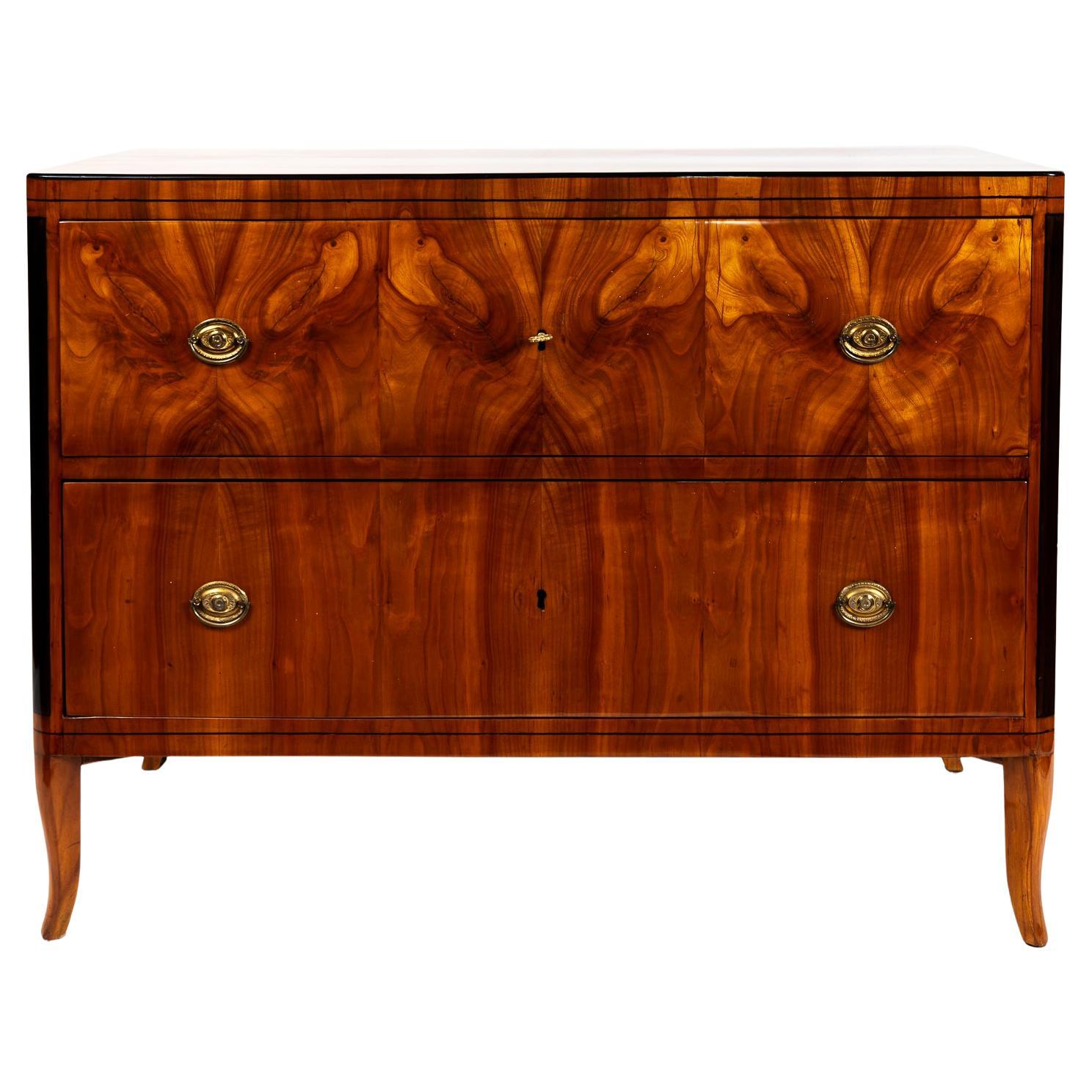 A most lovely early Biedermeier deep two-drawer chest on tall splayed legs made in a striking blonde cherry wood matchbook veneer on pine with ebonized black detailing. Each drawer opening with oval shaped brass pulls and a working key.  Note the