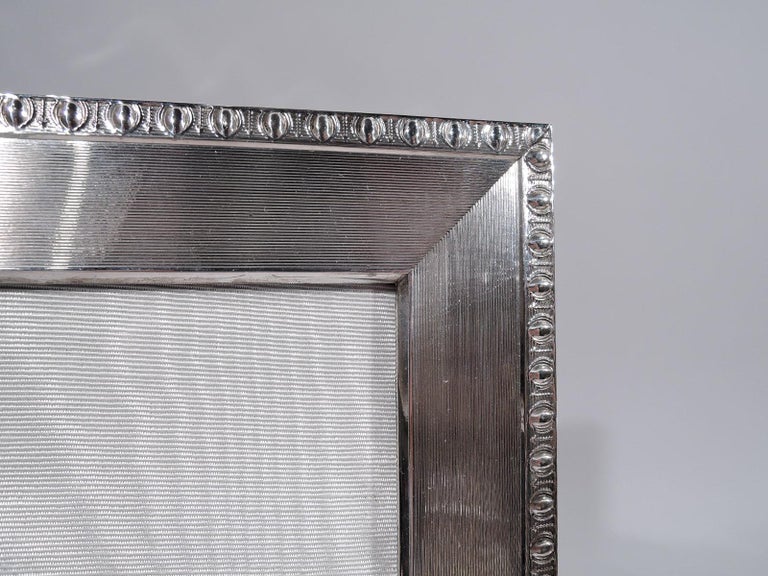 German midcentury Classical 800 silver picture frame. Rectangular window in flat surround with wraparound engine-turned lines, and raised egg-and-dart border. With glass, silk lining, and wood laminate back with hinged easel support for portrait