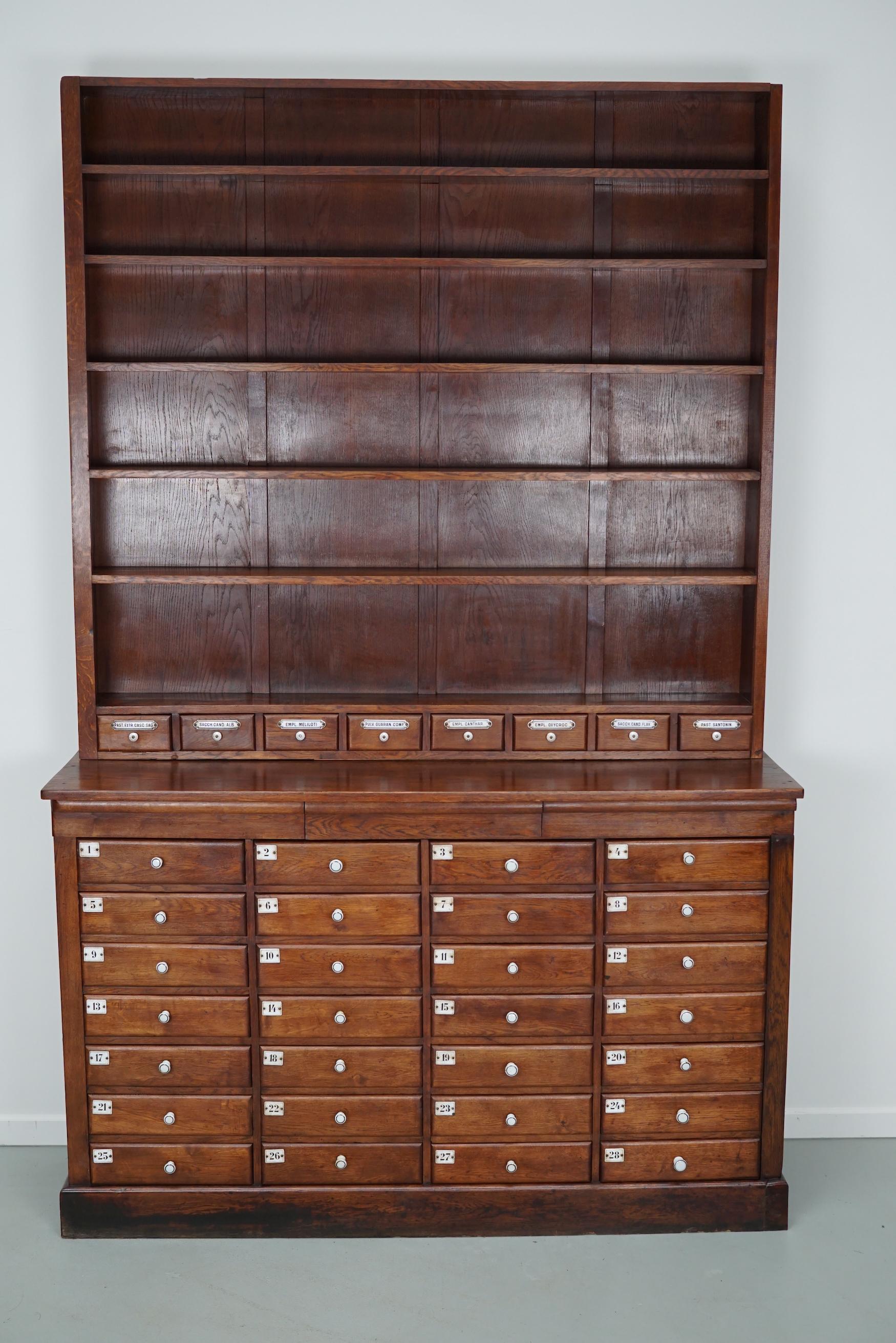 This apothecary cabinet was designed and made in the late 19th century in Germany. It is made from oak with porcelain knobs and enamel name and number plates. The cabinet has retained a rich patina with some distressing and signs of old repairs. The