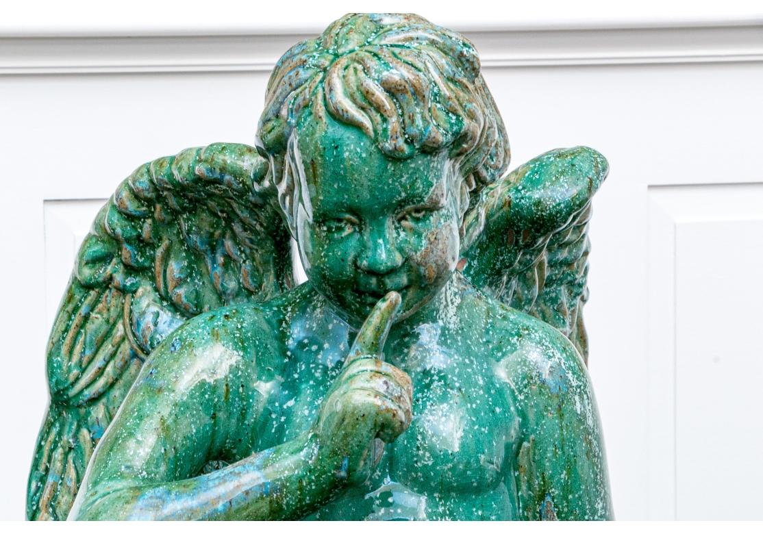 Une grande sculpture de jardin sous la forme d'un putto ailé assis, la main aux lèvres, semblant faire taire. Le putto est assis sur une plinthe au milieu des fleurs et a une main sur un carquois. La glaçure verte est entremêlée de taches d'autres