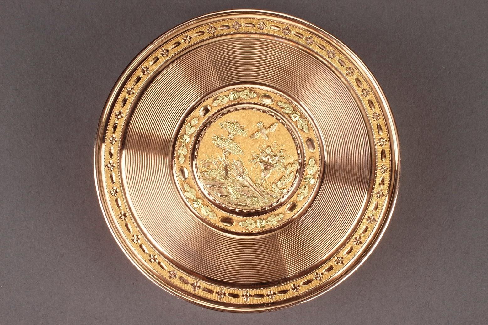 Große, runde Dose mit zwei Goldtönen. Der abnehmbare Deckel ist mit detaillierten, konzentrischen Linien und einem zentralen Medaillon verziert, das eine Taube darstellt, die über einen Blumenkorb und einen Köcher auf Amati-Goldgrund fliegt. Der