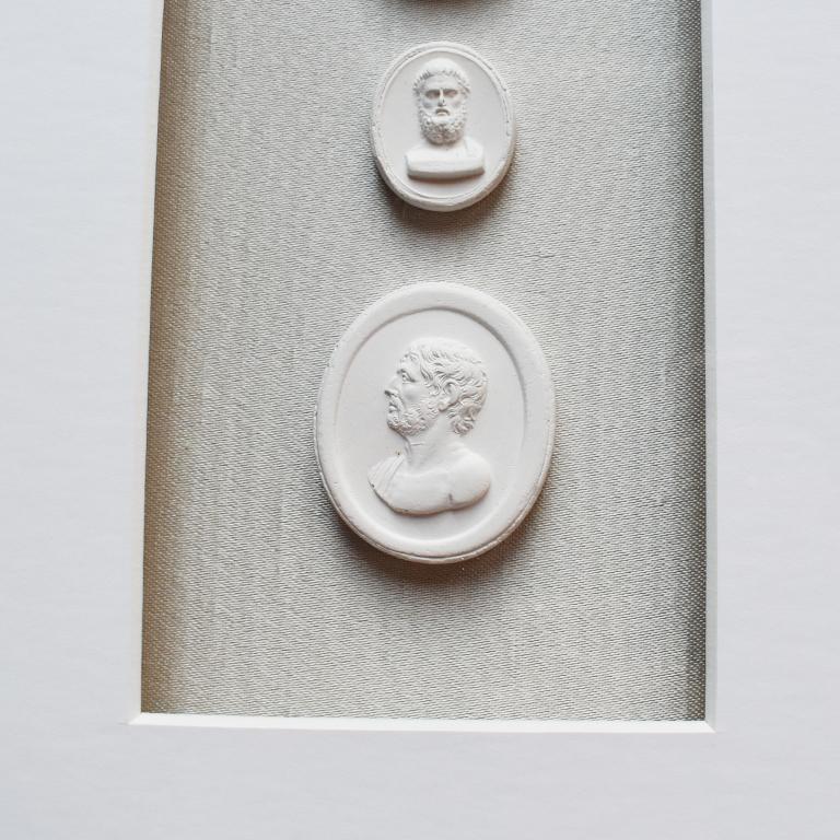 Vertical Italian collection of three framed intaglios on light gray silk background. This oversized portrait piece is framed in a gold rectangular frame. Inside, are three oval-shaped cameos of plaster. Each cameo is of a man with a beard. (We call