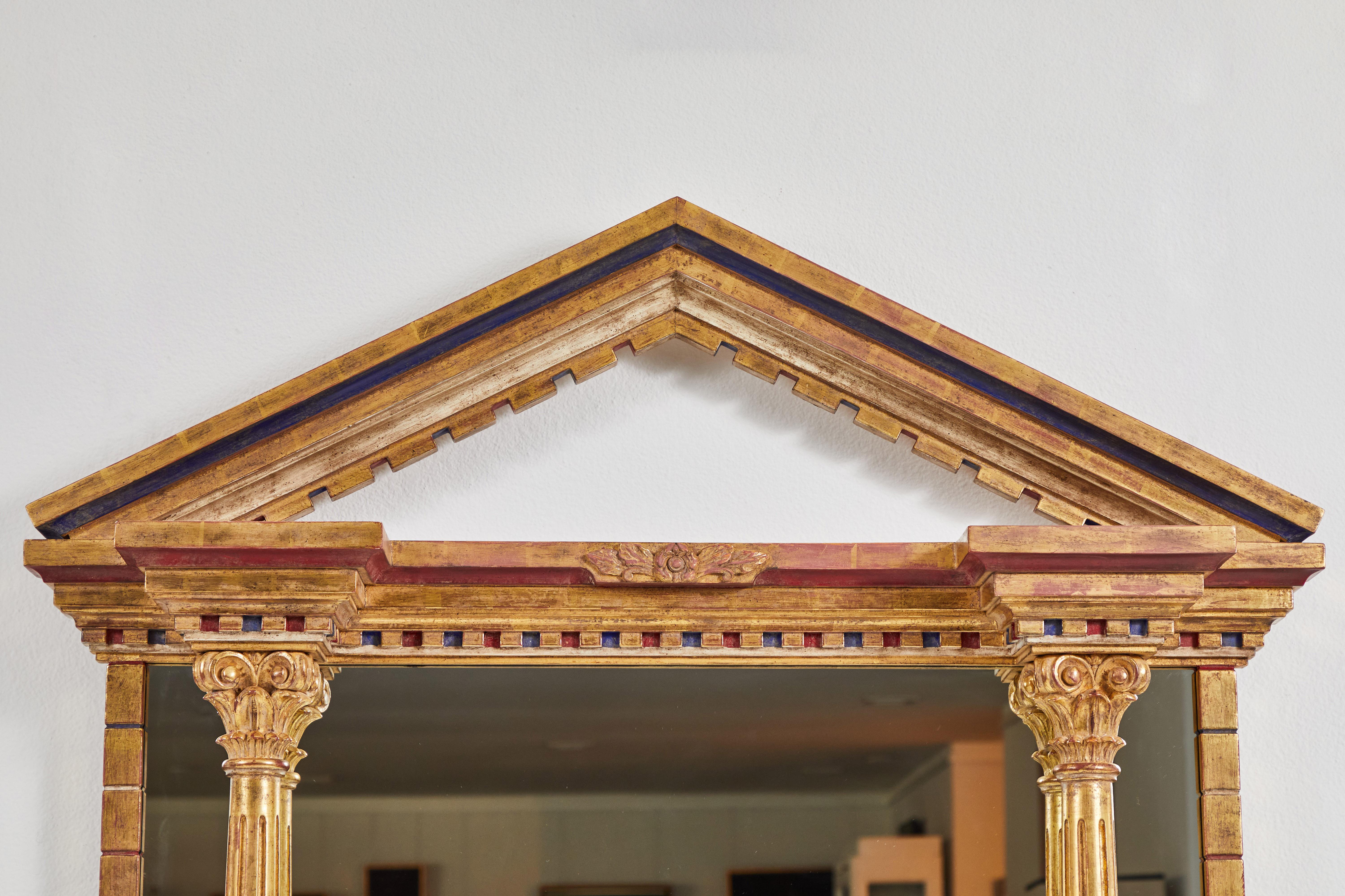 This grand gold leaf polychromed mirror boasts a decorated pediment reaching 63 inches high. Its carved indentations have been hand painted red and blue. The two columns on either side finish with a lotus. The mirror itself measures 39