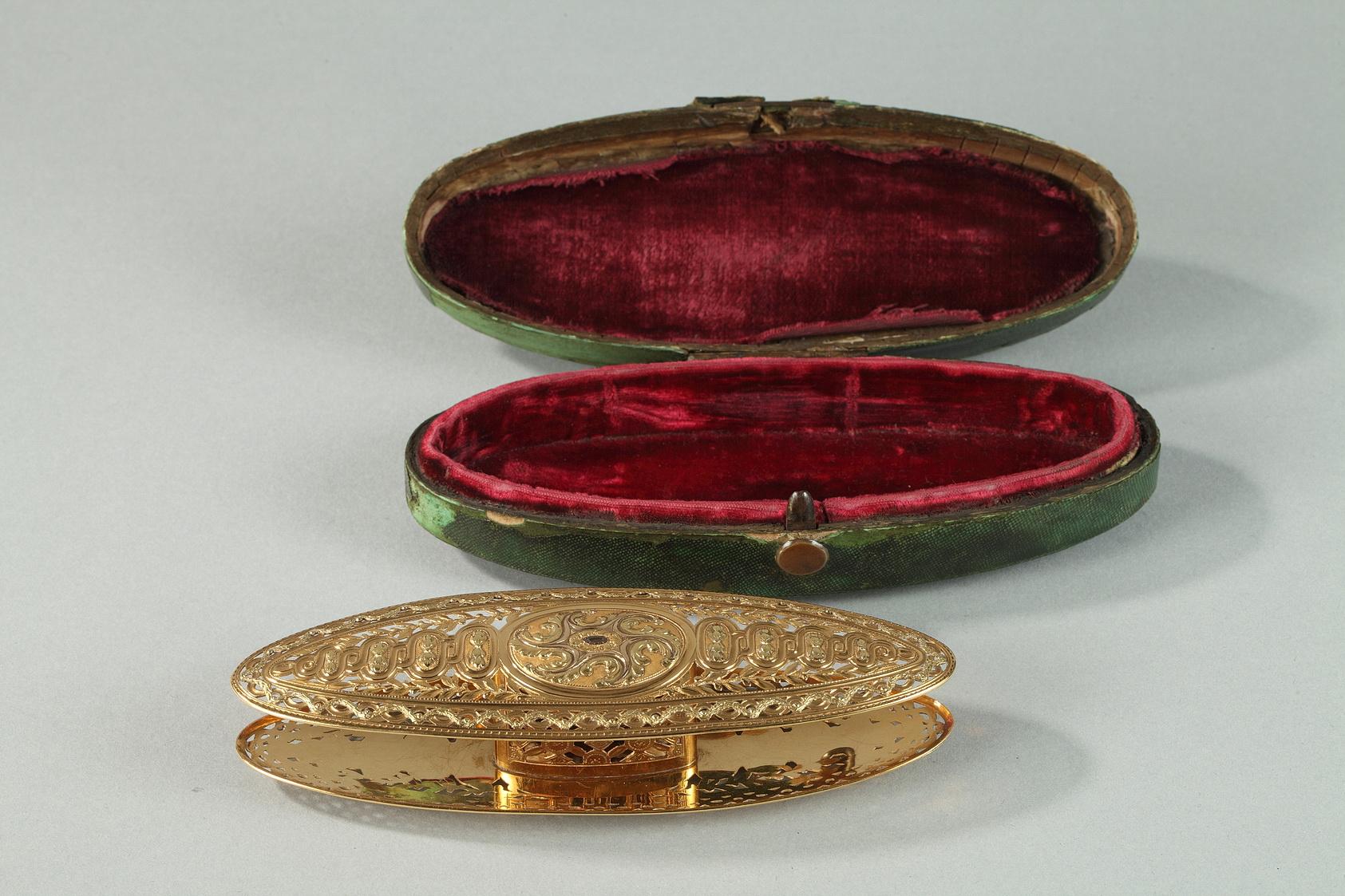 Large spool in colored gold. It is embellished with openwork laurel garlands and a row of gradated ovals running down the center with a stylized floral motif in the center of each oval. A large, central oval is decorated with white gold crown