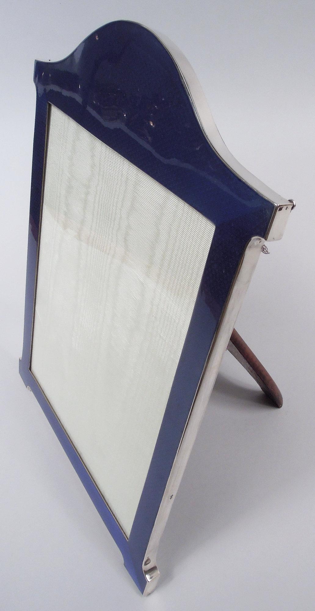 Portarretratos Jorge V de plata de ley y esmalte. Fabricado por Sanders & Mackenzie en Birmingham en 1927. Ventana rectangular en marco con remate arqueado y soportes de ménsula. Esmalte guilloché azul en el anverso y plateado en los laterales. Gran