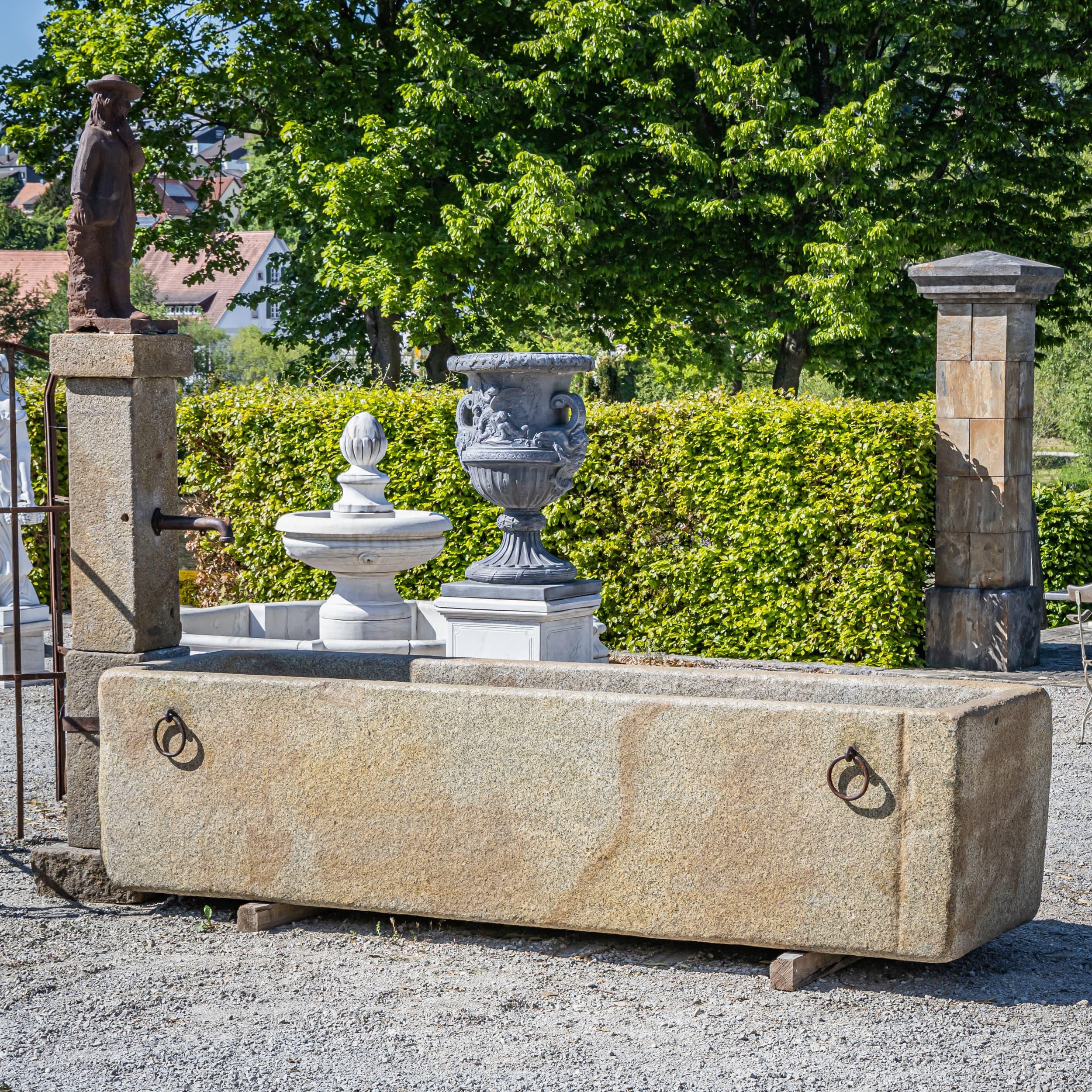 Striking fountain made of solid, hand-hewn granite. The rectangular basin measures 90 × 370 × 120 cm (H × W × D), accompanied by a prominent spout column measuring 235 × 62 × 50 cm. With its rustic form and substantial craftsmanship, this piece