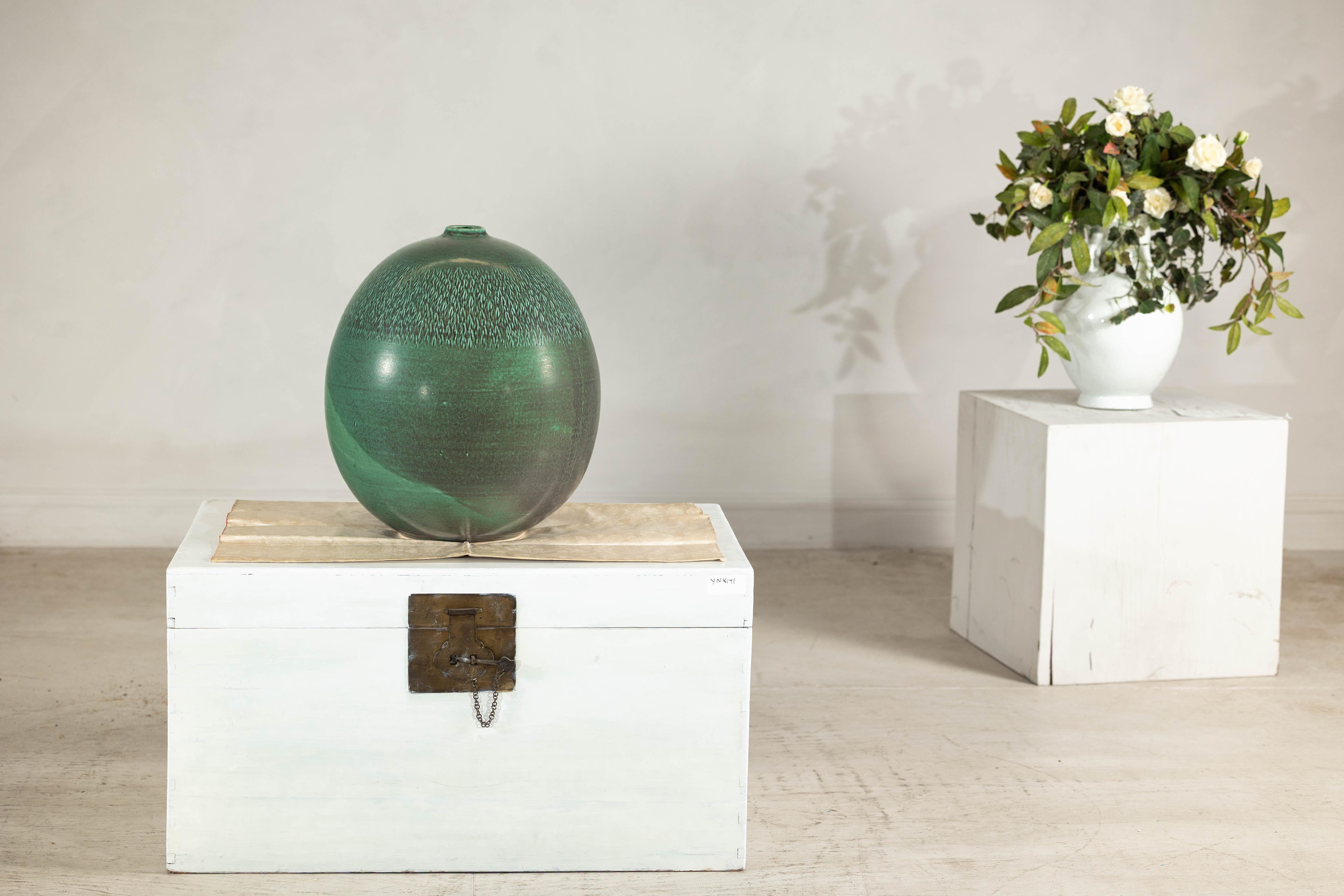 Étude de texture et de tonalité, ce vase artisanal fait une déclaration élégante avec sa glaçure vert profond et ses détails de surface complexes. La forme sphérique est adoucie par une finition satinée, tandis que le motif au sommet crée une
