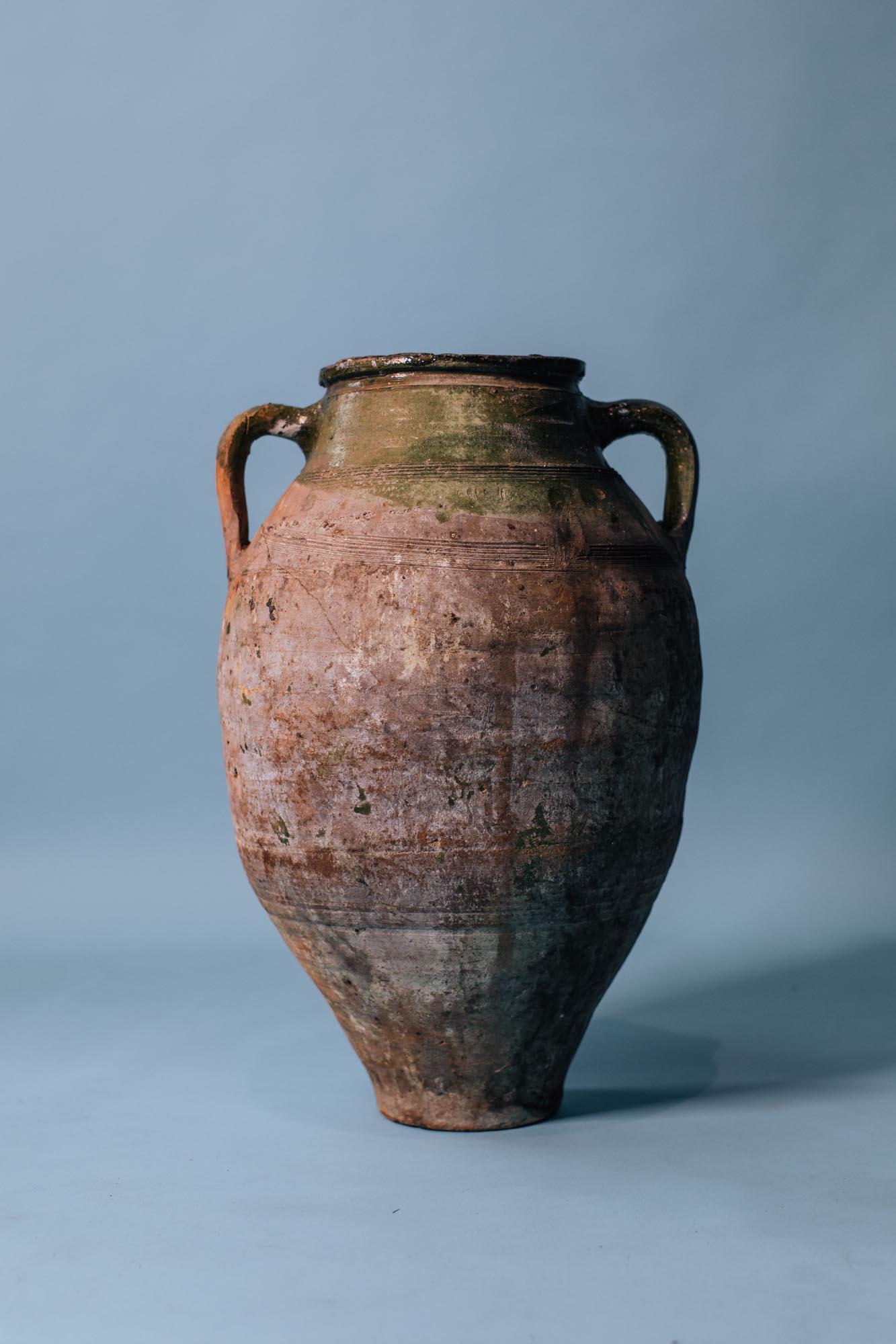 A striking early 20th-century terracotta storage jar, hand-thrown in Anatolia and finished with a soft olive-green glaze that gently fades into natural clay. Once used to store olive oil and grains, it carries the hallmarks of traditional