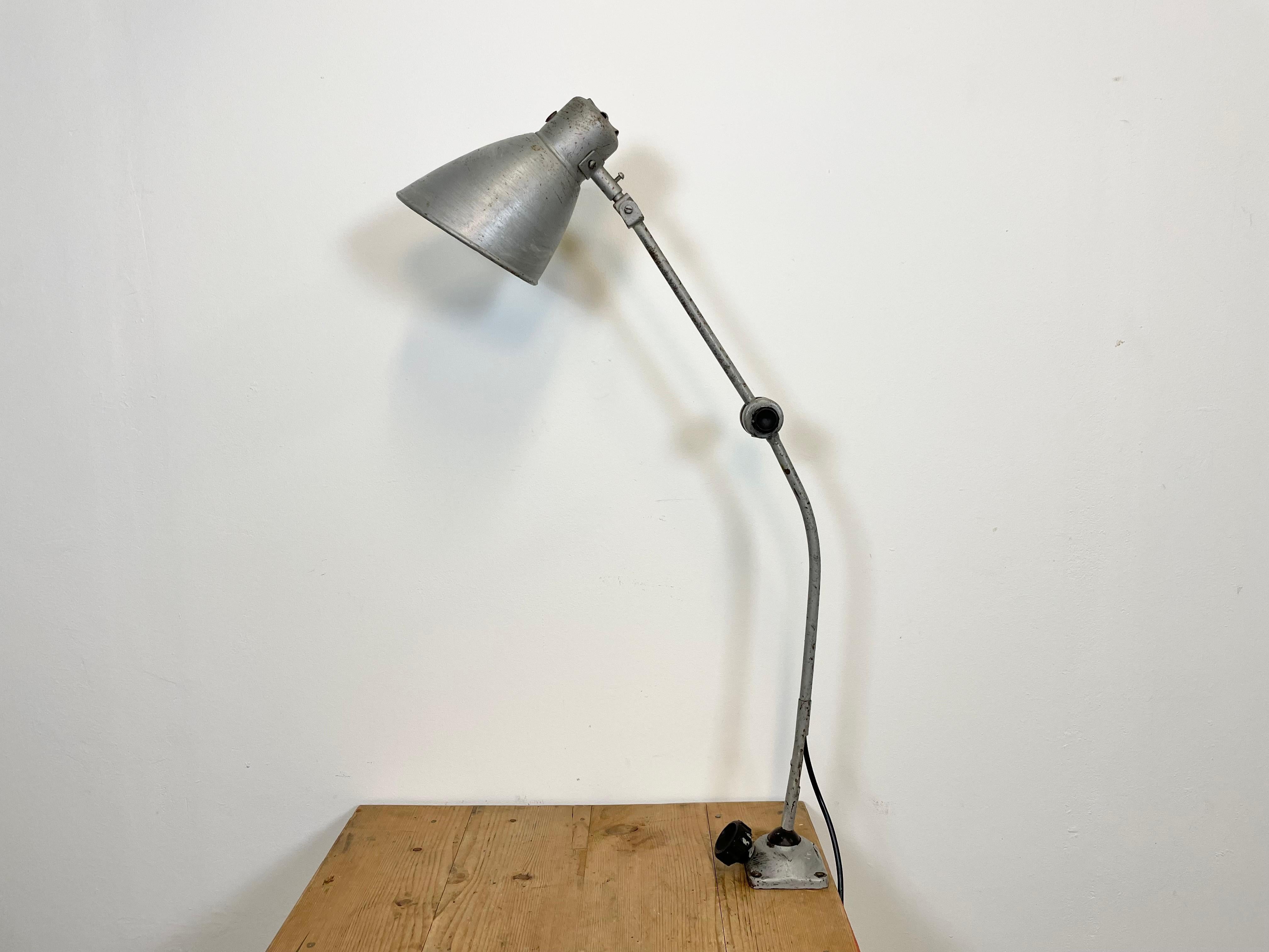 Lampada da tavolo industriale di grandi dimensioni con tre snodi regolabili realizzata nell'ex Cecoslovacchia negli anni '60. Presenta una base, un braccio e un paralume in ferro grigio. L'attacco in porcellana richiede lampadine E 27/E 26. Un nuovo