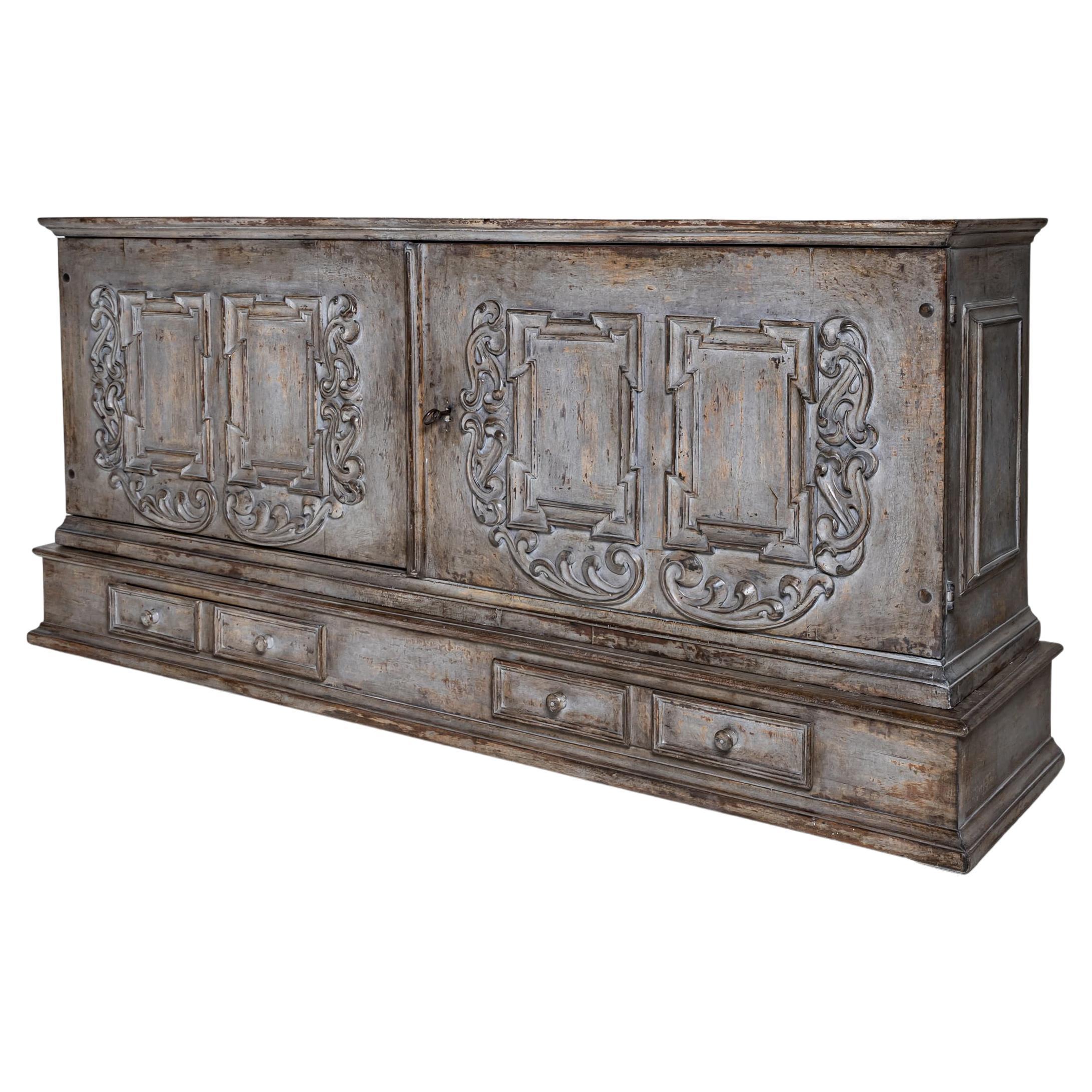 Impressive handcrafted sideboard from the 18th century, featuring two doors with richly carved decorative elements. The gray finish enhances the intricate, symmetrical relief motifs. A continuous row of drawers is integrated into the lower section.