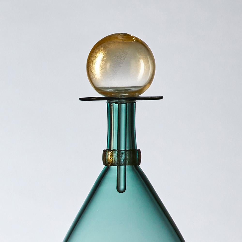 Contemporary hand blown glass bottle in a graceful eliipse form, inspired by apothecary decanters of Mid-Century Modern design. Coordinated neck-wrap and blown glass stopper are finished with gold-leaf. Offered in Tourmaline, a smoky blue-green,