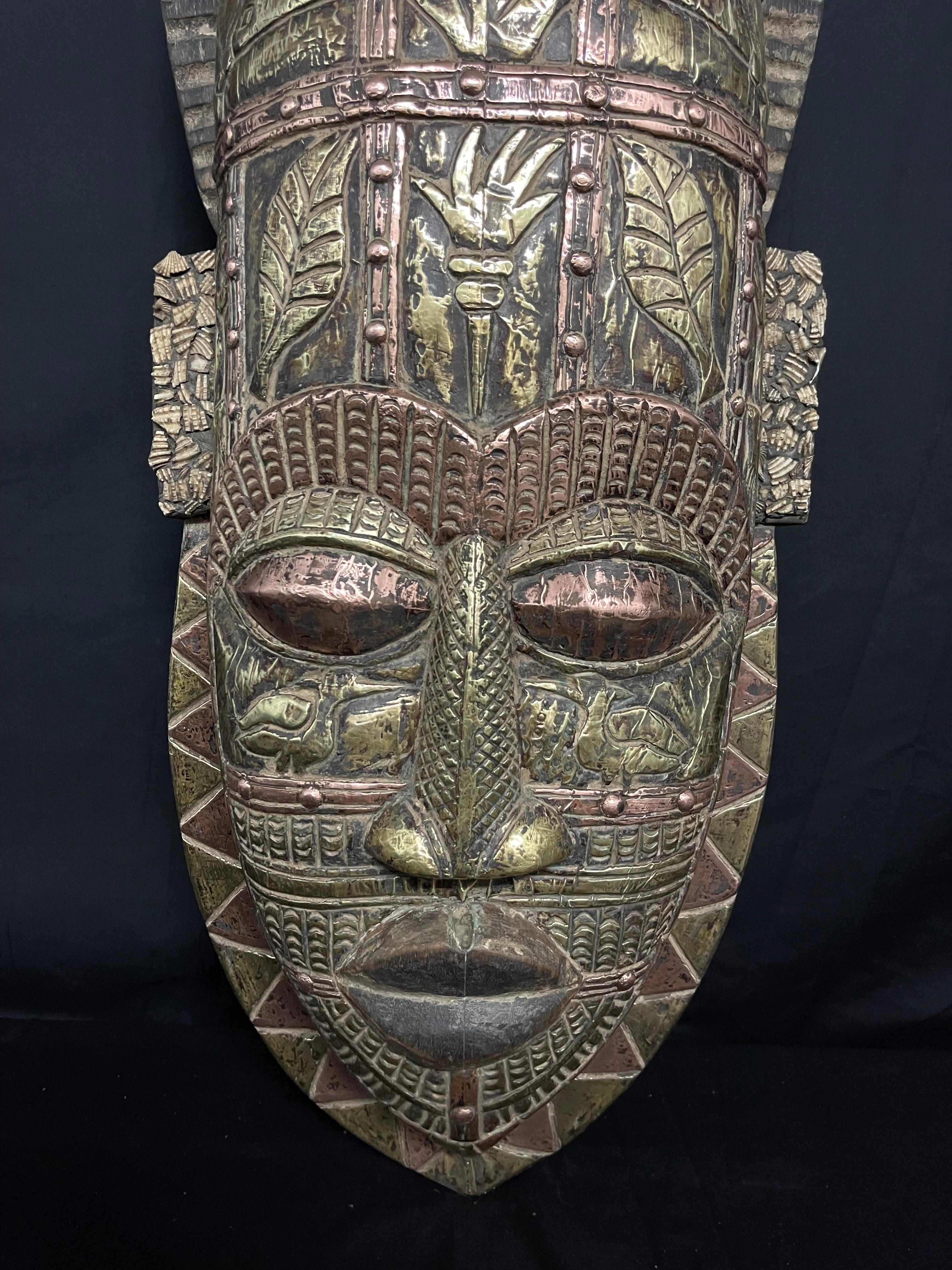 Esta gran máscara tribal africana tallada a mano muestra una artesanía intrincada con sus detalles de cobre batido y hojalata, realzados por vibrantes acentos de mosaico de conchas. Estas máscaras suelen tener significados culturales importantes y