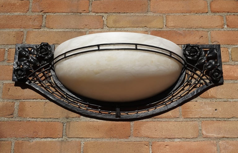 Impressive and sizable wall sconce with a perfect alabaster shade.

This rare Art Deco style sconce is beautiful both in design and execution. The combination of the blackened iron with the handcrafted rose motifs on the one hand and the creamy