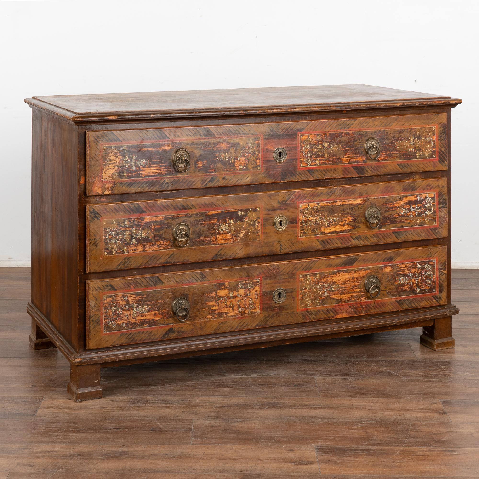 The soft colors are all original on this hand-painted antique chest of 3 drawers from Hungary.
The lovely panels with hand painted flourishes were a traditional folk art design of the era set against a brown faux wood finish, adding a cheerful feel
