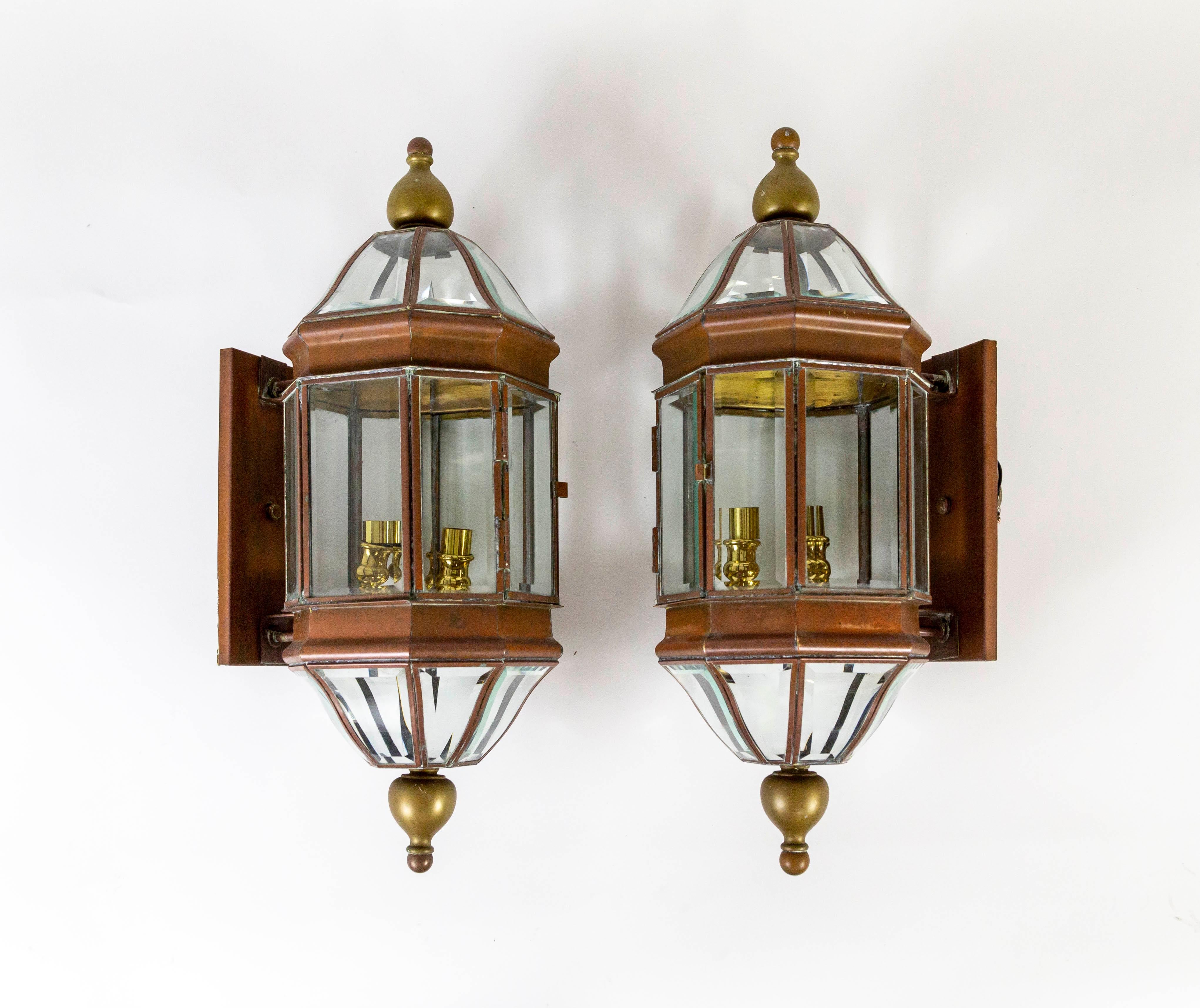 A pair of sizable, 3-candlestick light, copper, and brass lantern sconces in an elongated, octagon shape curving at the top and bottom and finished with finials. The segmented glass panels are beveled, held in a copper frame slightly projected from