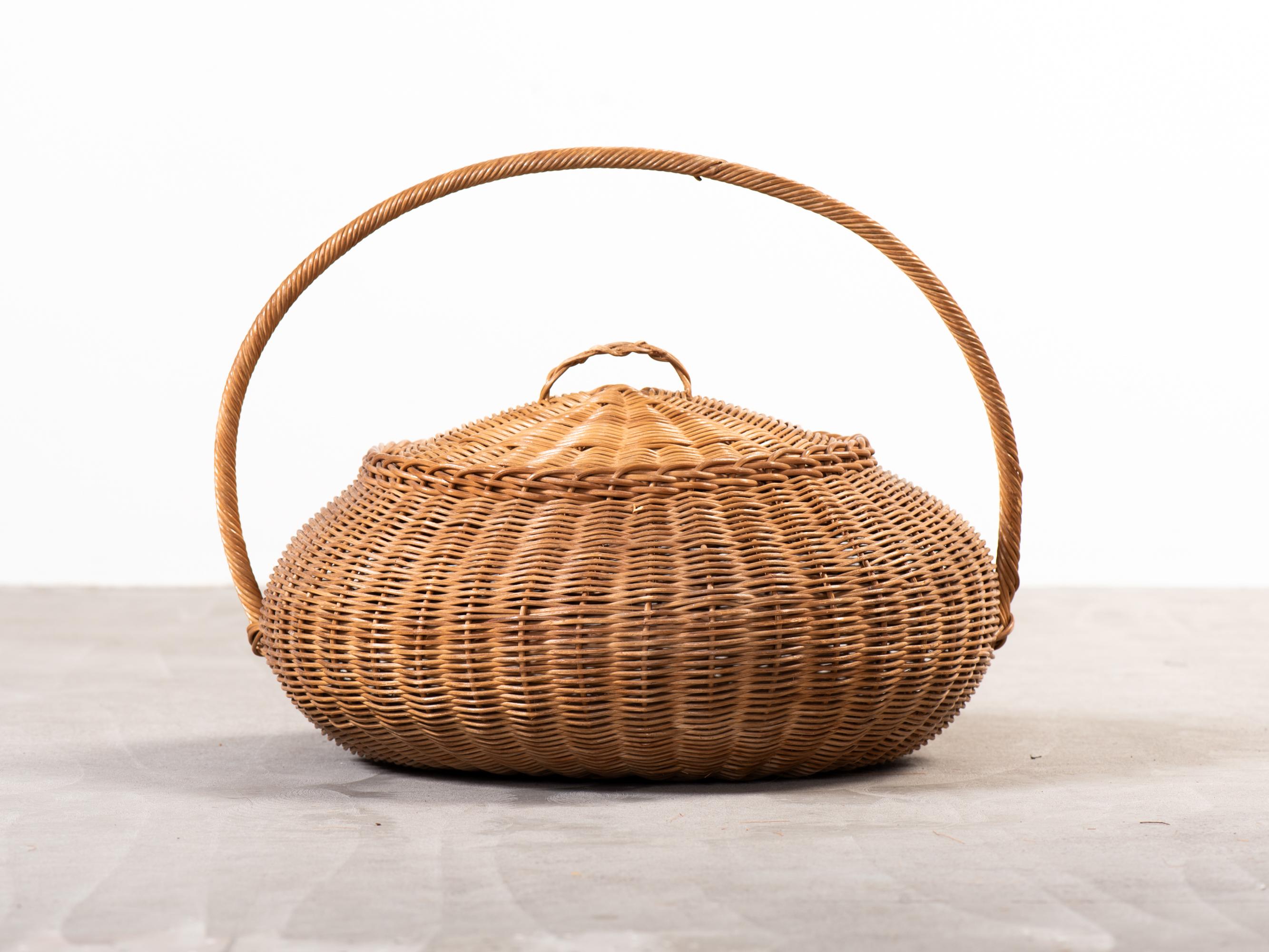 Una gran cesta de tapa redondeada, con un perfil suavemente abombado y un asa escultural envuelta en una torsión apretada de caña. Tejido a mano con ratán partido, el dibujo de la superficie es sutil y uniforme, con un fino ritmo vertical y un