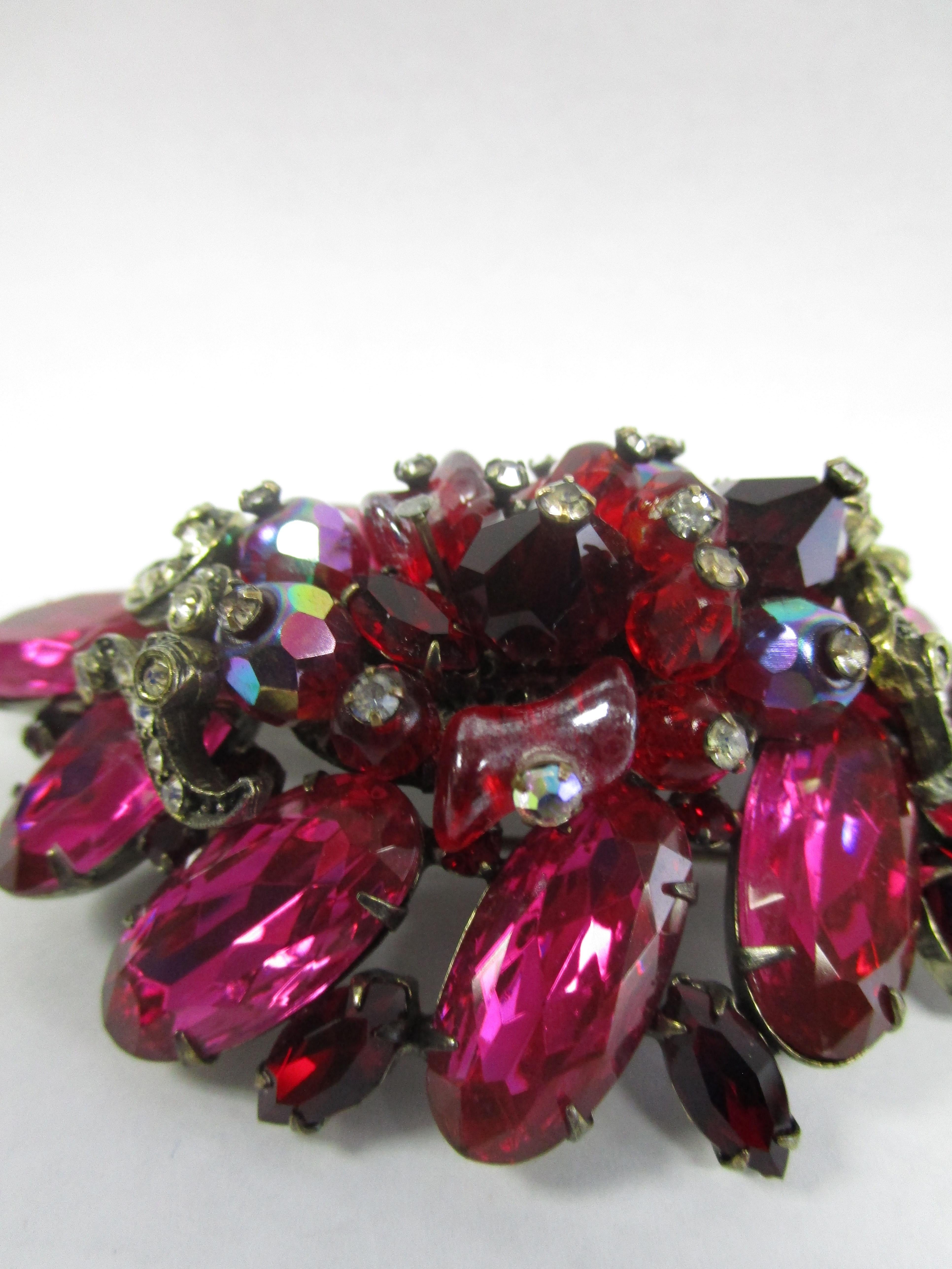 Hattie Carnegie Pink, Red, and Clear Bead and Glass Brooch. We present to you this beautiful and romantic berry colored glass brooch from Hattie Carnegie! It features an array of reds, pinks, purples and clear cut stones and faceted beads. Red glass