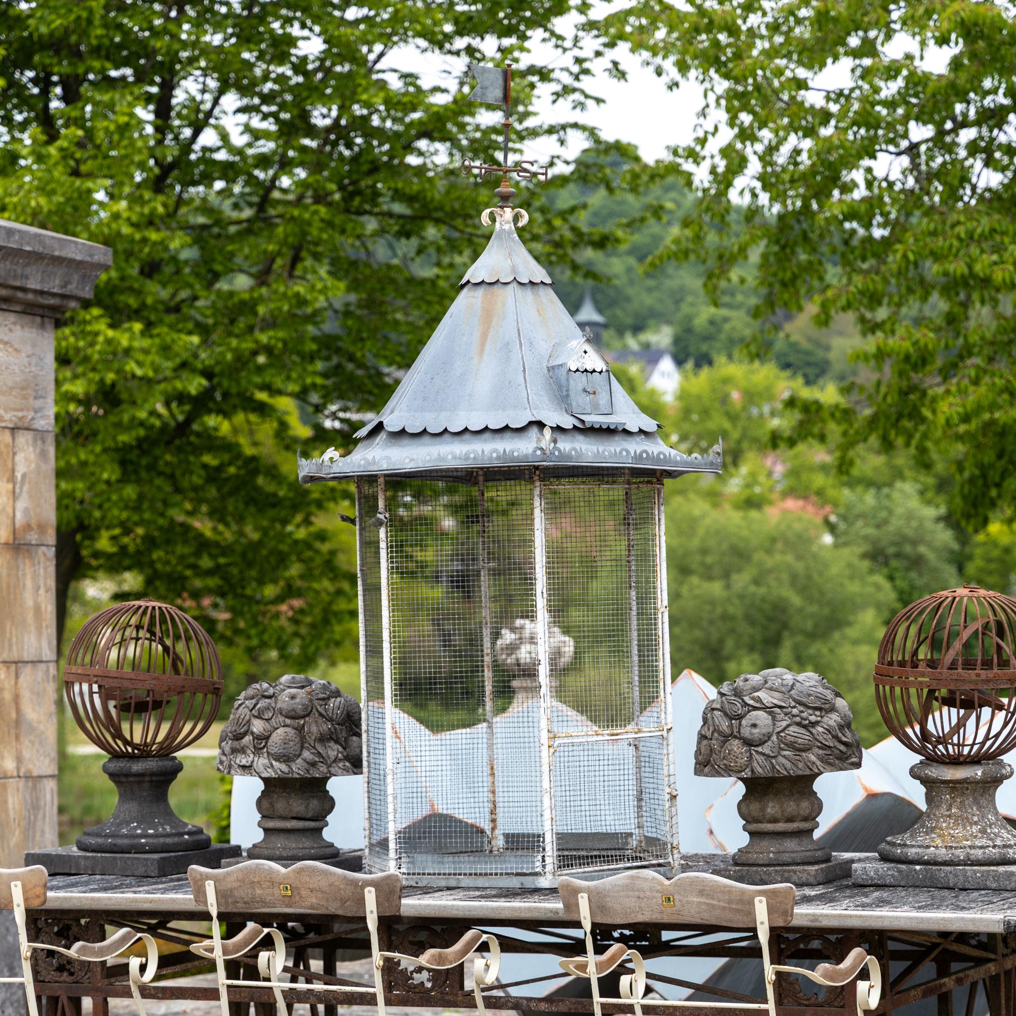 Decorative hexagonal birdcage with a pointed roof and scalloped metal edge, dating to the late 19th century. The top is crowned by a crab and a weather vane. The lower section is painted white, and the upper part is enclosed in fine metal mesh.