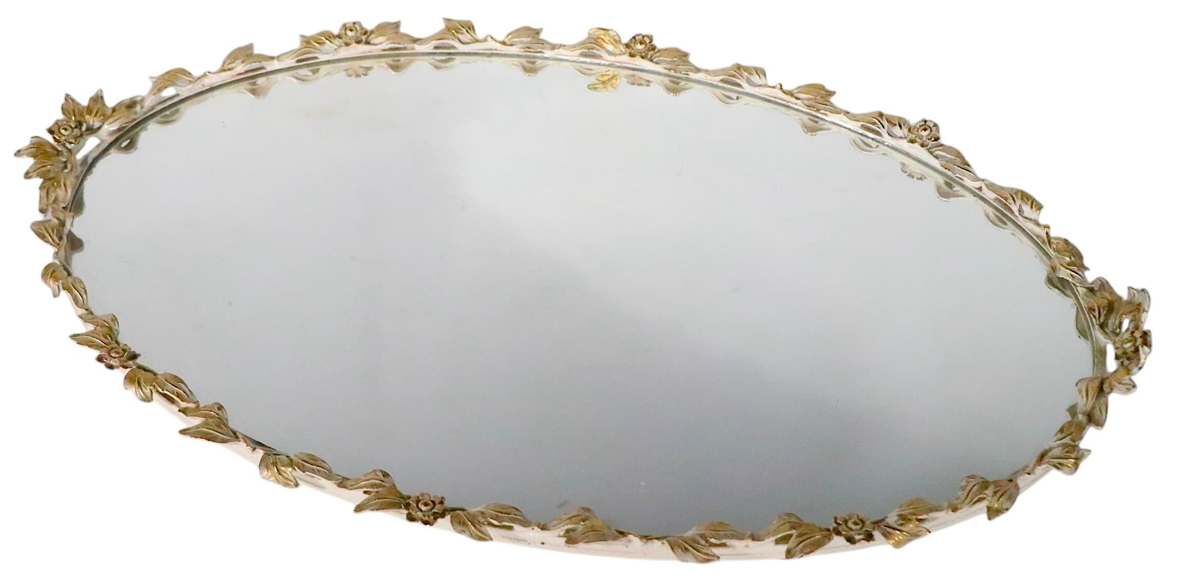 Exquisite oval vanity, decorative table, or barware, tray having a cast brass frame with original mirror surface. The tray features  decorative metal foliate trim,  with leaves and flowers, it is in original two tone white paint  and brass finish.