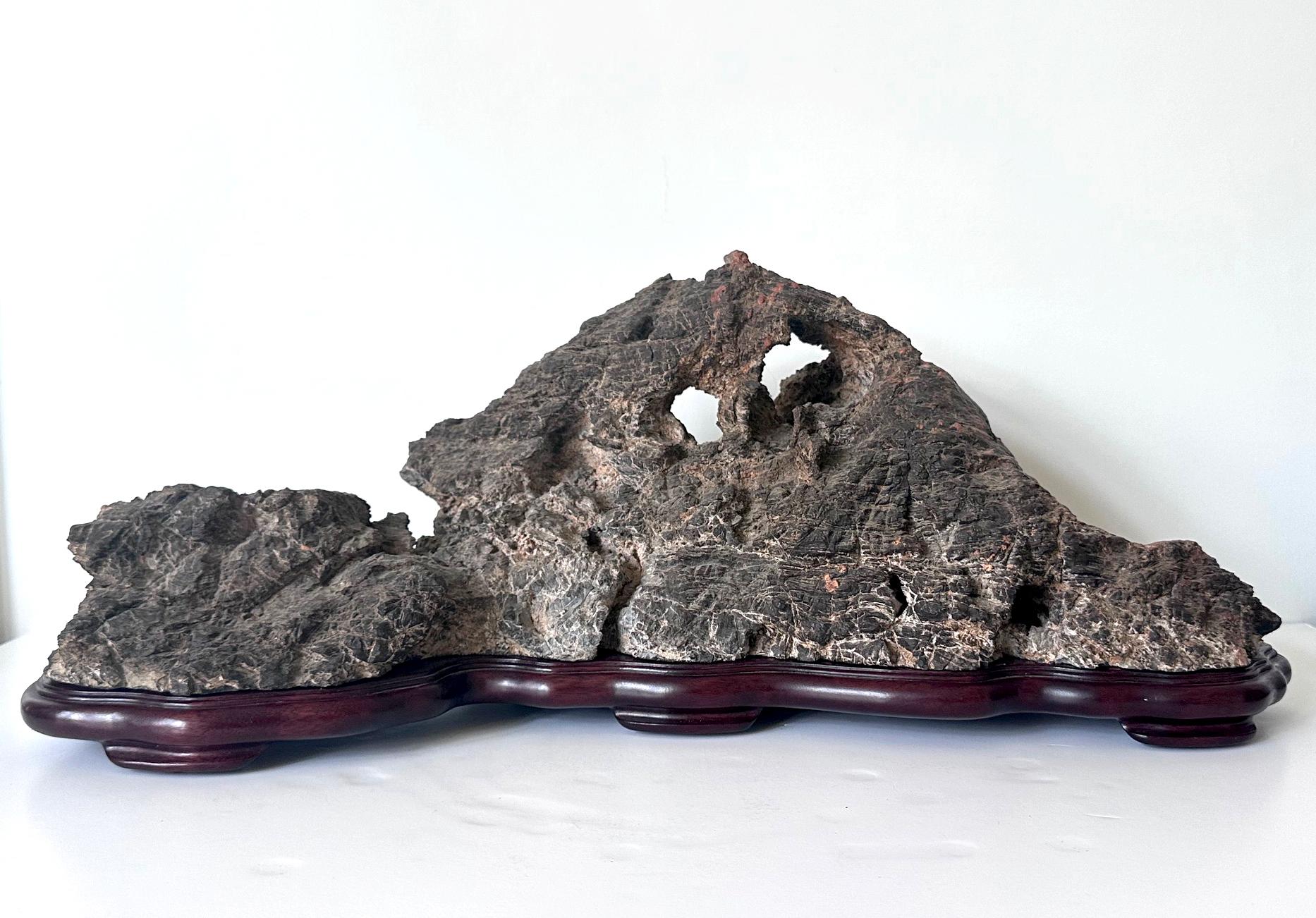 Pierre de lettré chinoise de type Lingbi de forme horizontale présentée sur une base en bois sculpté conforme. Impressionnante par sa taille et sa forme, cette pierre de Lingbi se manifeste par une silouette de chaîne de montagnes. La surface