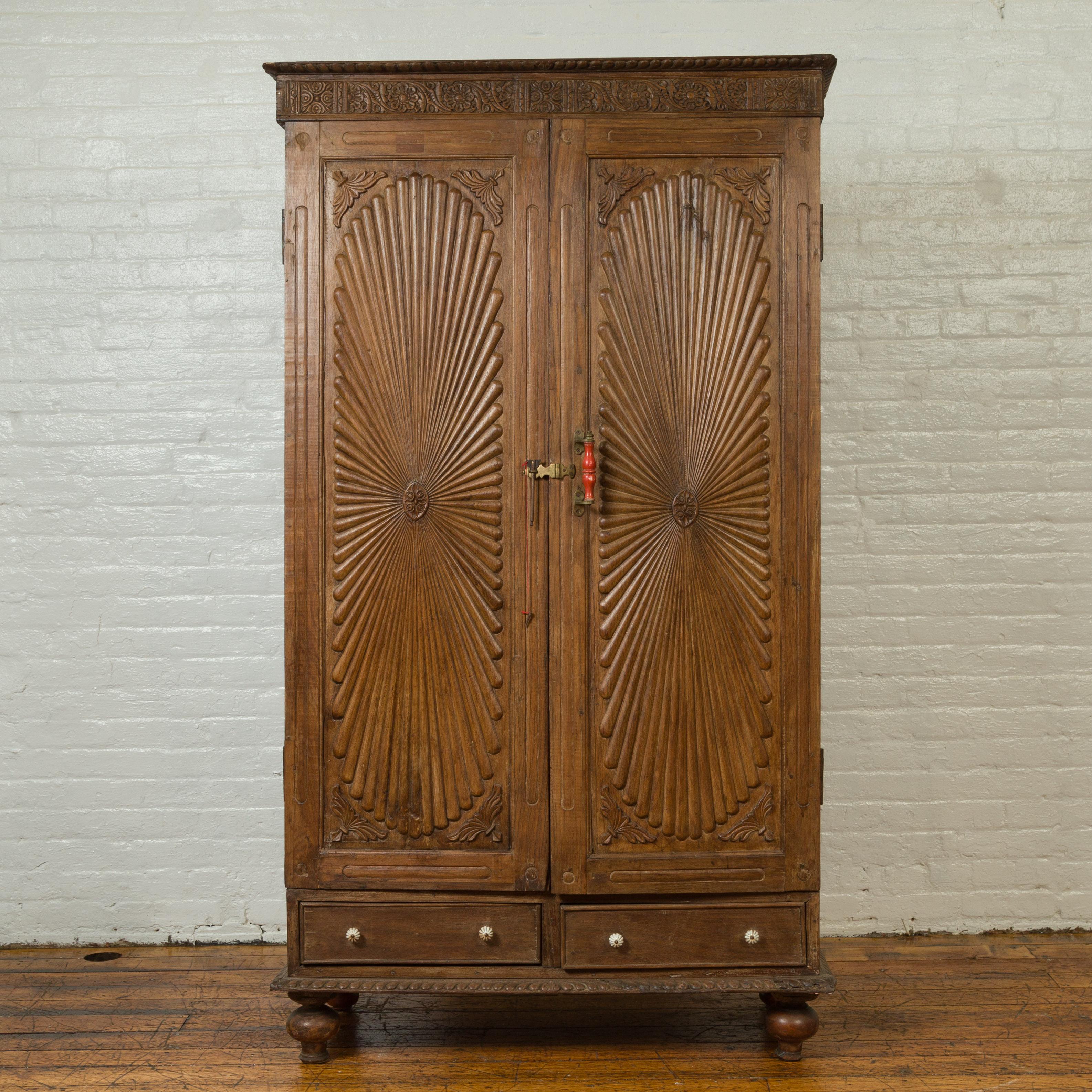 An antique Indian wooden cabinet from the 19th century, with sunburst patterns and floral motifs. Born in India during the 19th century, this tall cabinet features a carved cornice adorned with floral motifs, sitting above two doors presenting large