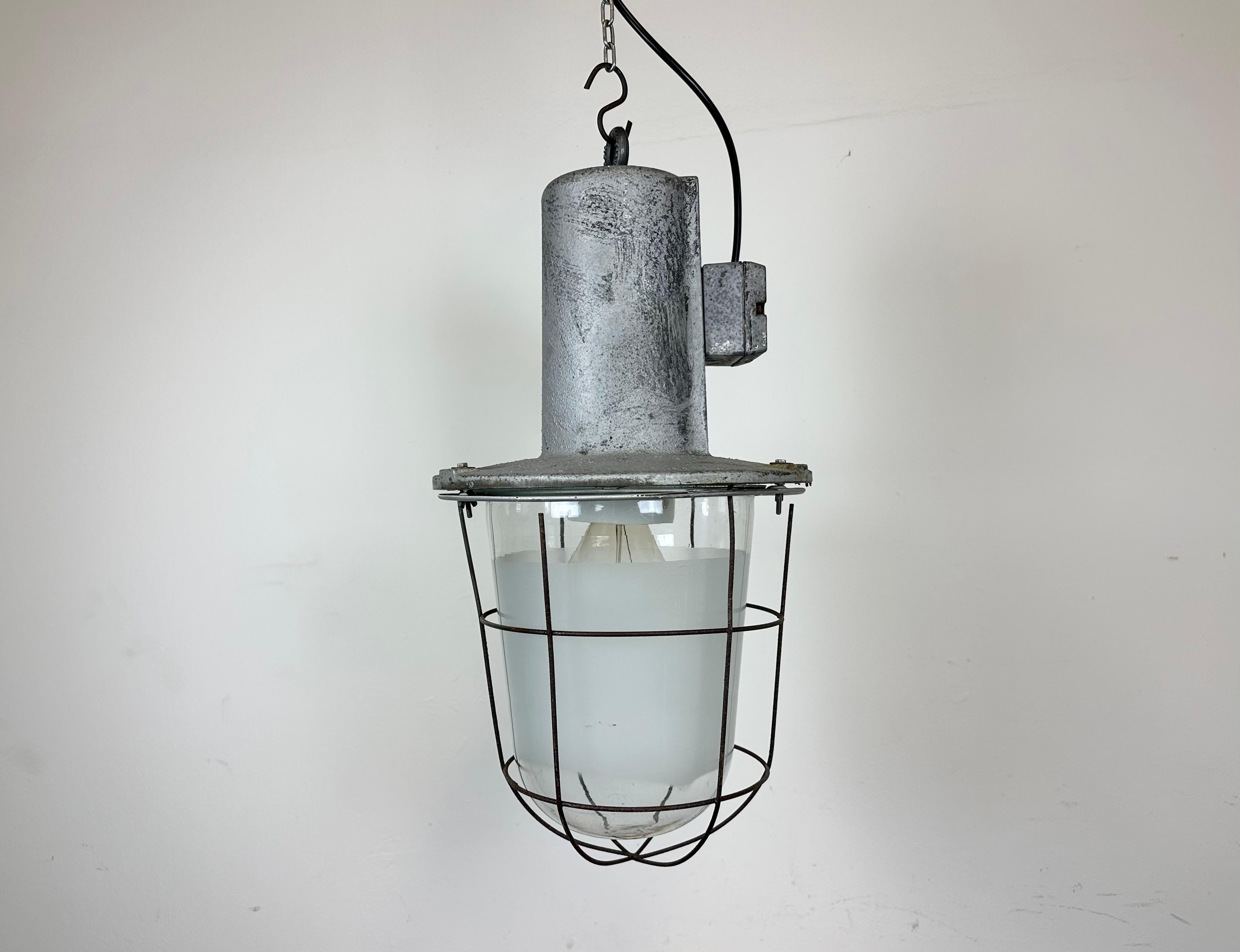 Large industrial factory hanging lamp made in former Czechoslovakia during the 1960s. It features a cast aluminium body, a partly clear glass and partly milk glass cover and an iron grid. The porcelain socket requires standard E 27/ E 26 lightbulbs.