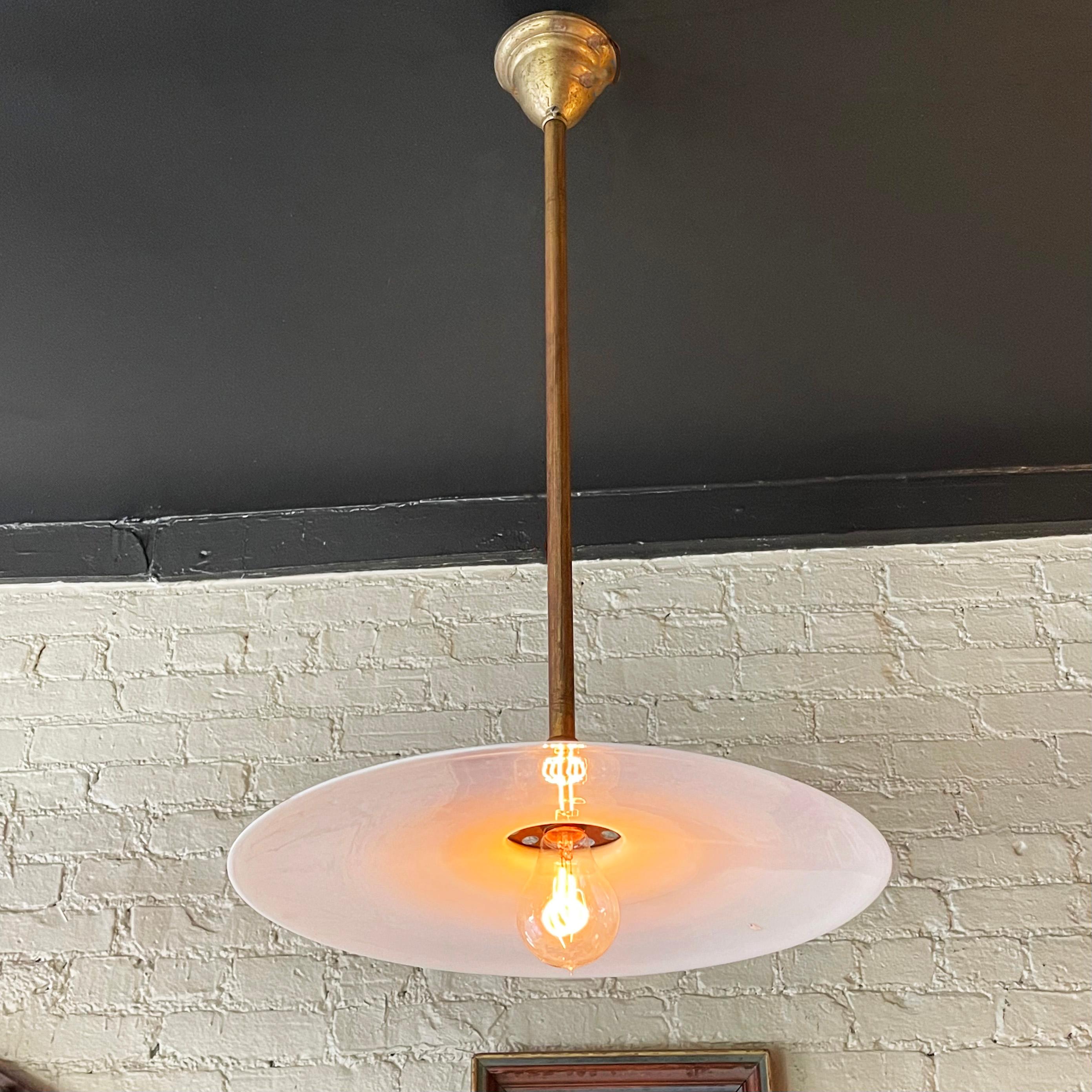 Early 20th century, industrial pendant light features a large, opaque, milk glass disc shade on a patinated brass pole with brass canopy and paddle switch hardware.