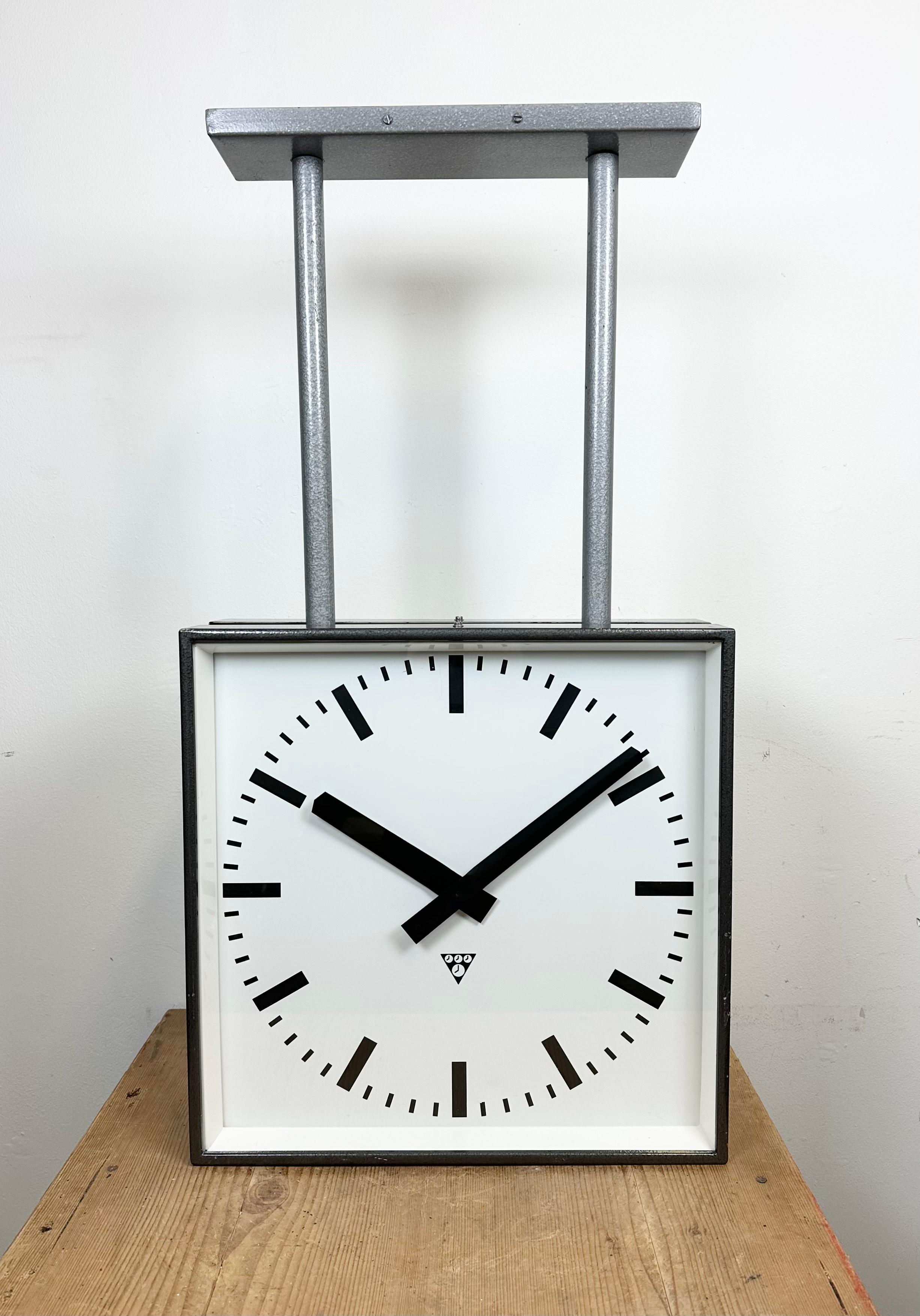 This square double-sided railway, school or factory wall clock was produced by Pragotron, in former Czechoslovakia, during the 1970s. The piece features a grey metal body, a dark grey hammerpaint clock frames, a clear glass covers, and an aluminum