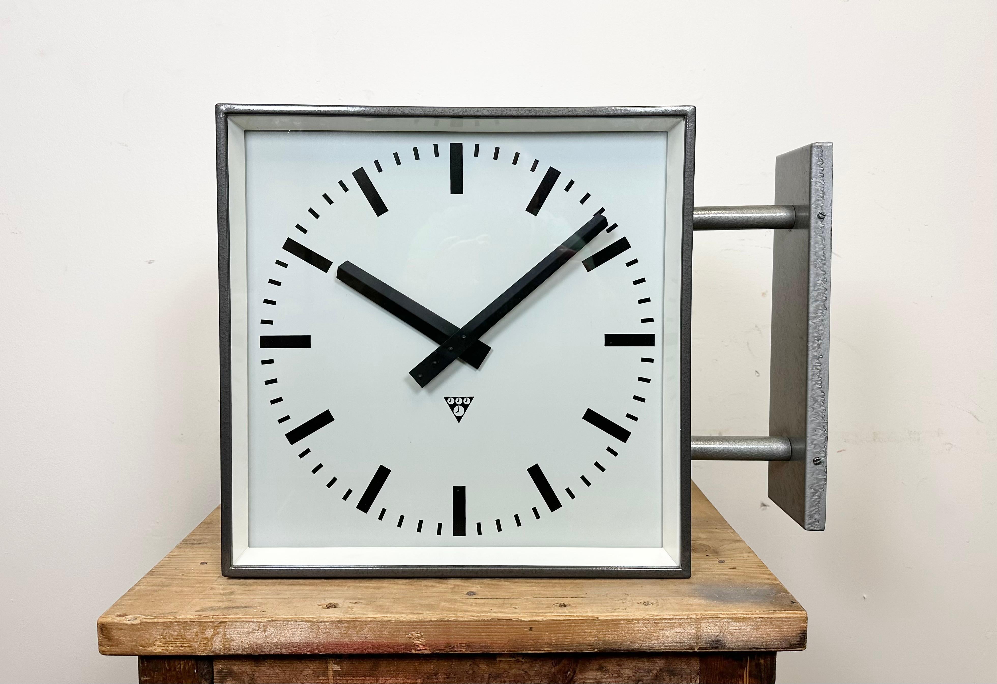 This square double sided railway, school or factory wall clock was produced by Pragotron, in former Czechoslovakia, during the 1970s. The piece features a grey metal body, a dark grey hammer paint clock frames, a clear glass covers, and an aluminum