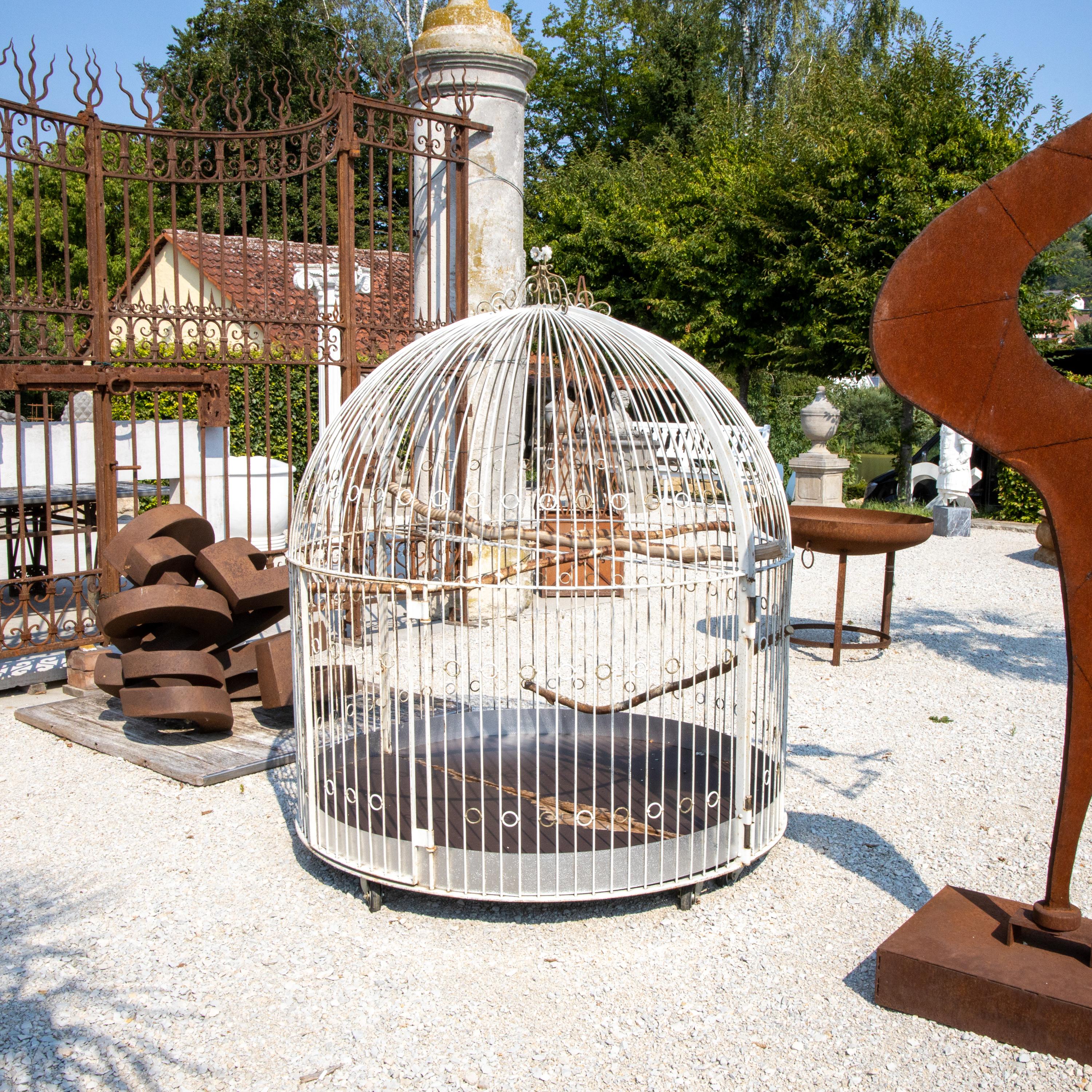 Großer Vogelkäfig oder Voliere aus weiß gestrichenen Eisenstäben mit Ringelementen und krönendem Blumendekor. Der Käfig steht auf Rädern und kann in der Mitte geteilt werden.
   