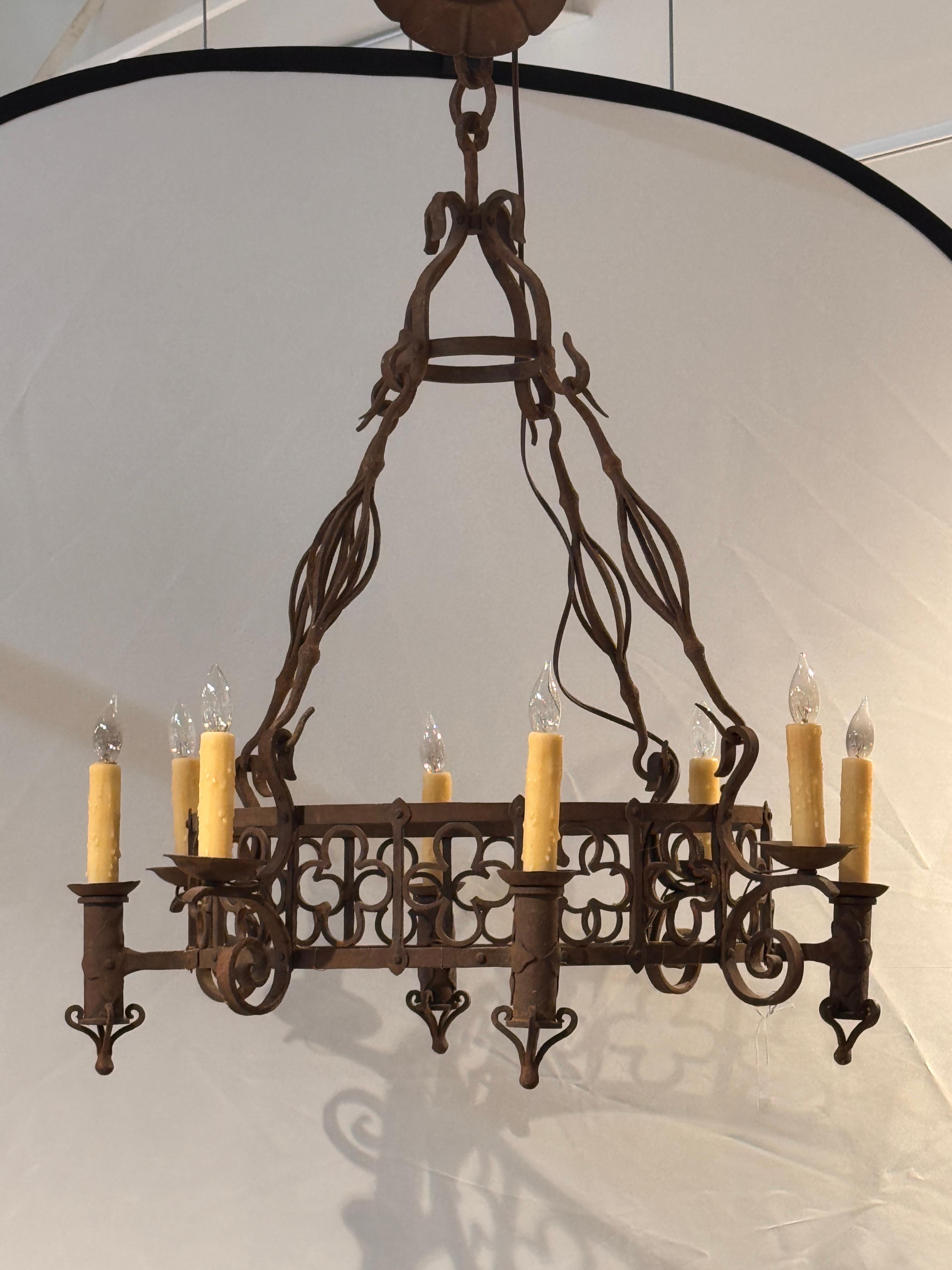 Substantial iron chandelier featuring eight lights and a rectangular frame with decorative quatrefoil and scroll motifs. The forged iron arms extend outward with curled supports, while the top is suspended by twisted iron straps. Candle-style