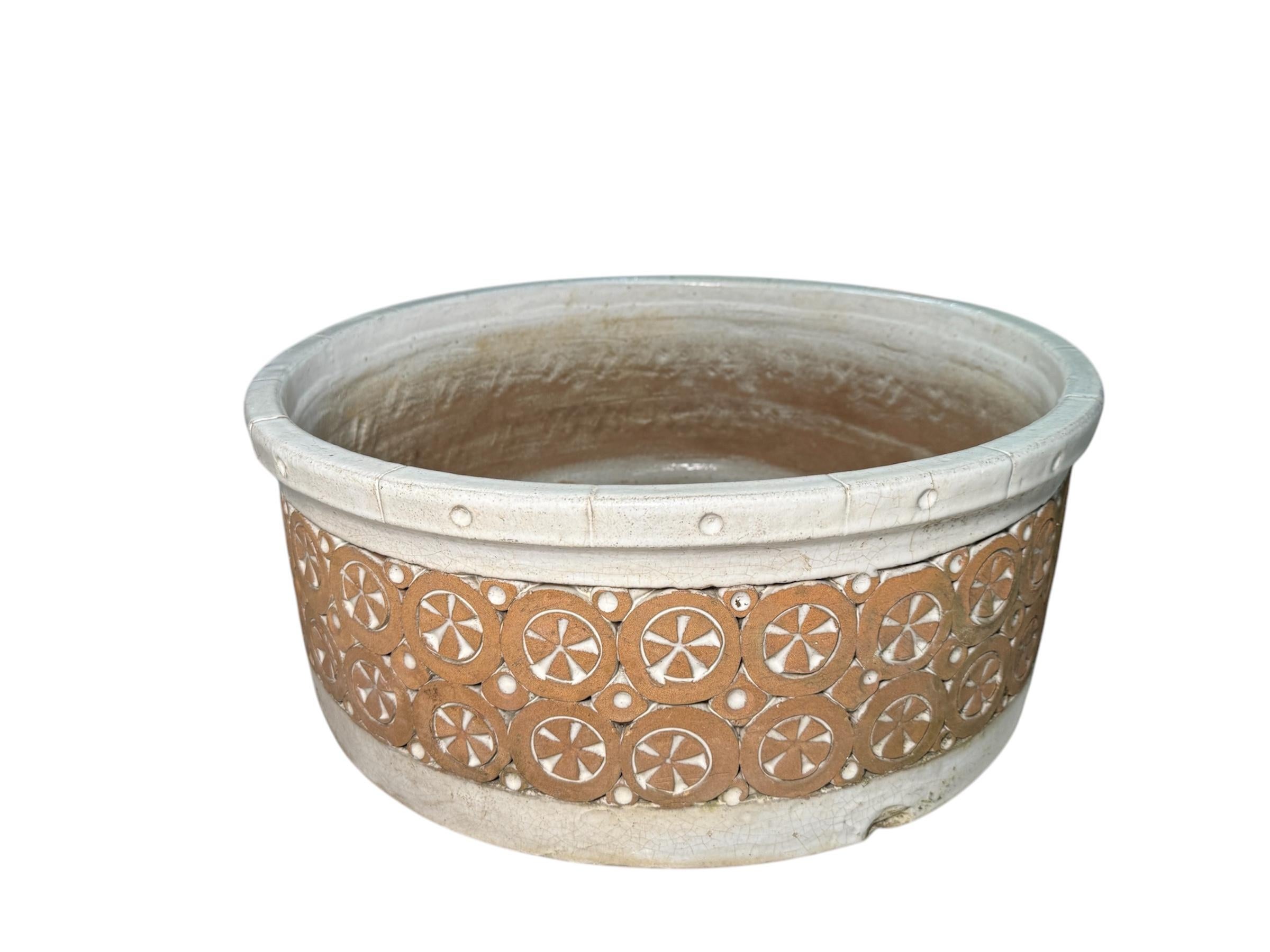 This is an Isla Del Sol Stoneware Bowl Planter. It depicts an oval white painted stoneware bowl decorated with two rows of circles adorned in the center with brick color flowers alternated with three rows of squares with concave hemispheric centers.